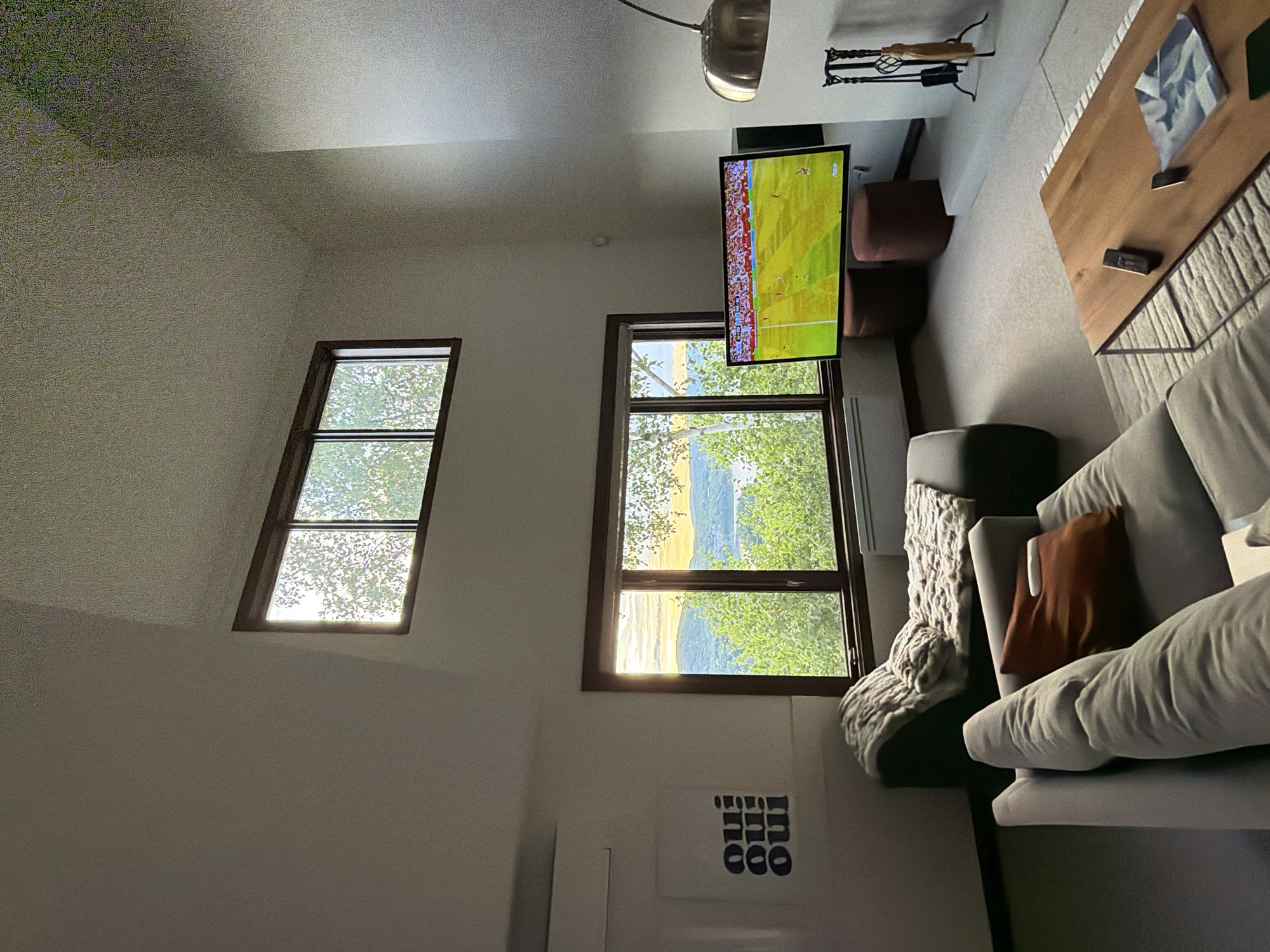 A living room in Mont-Tremblant, Canada, features a sectional sofa, a chaise lounge with a blanket, a large television displaying a soccer match, a coffee table with remotes and a magazine, and a fireplace set. Large windows offer a view of a body of water and trees, with the sun setting in the distance. A piece of art with the word "Oui" repeated is hung on the wall. The ceiling is high and angled, with a small window at the top. The room is well-lit by natural light and overhead lighting.