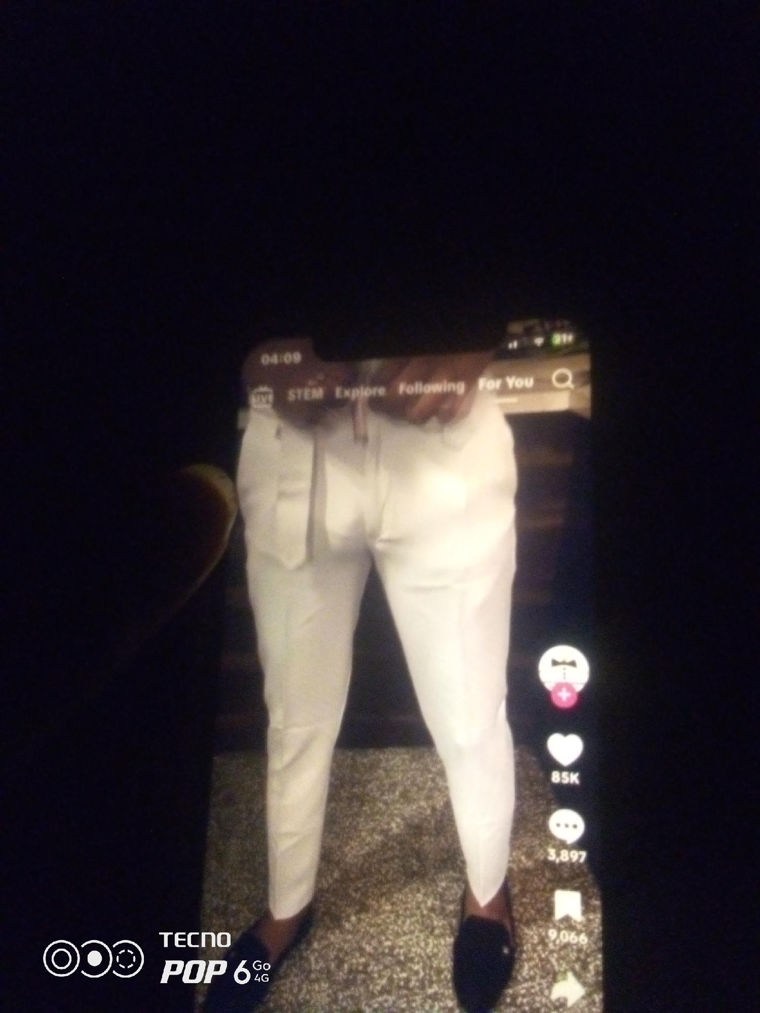 A person's lower body is visible, wearing white pants and dark shoes. The image appears to be a screen capture, possibly from a social media app, given the overlaid text and icons. The text reads "04:09," "LIVE," "STEM," "Explore," "Following," and "For You." On the right side of the screen, there are icons representing likes, comments, and shares, with numbers indicating counts: "85K," "3,897," and "9,066." The bottom left corner shows the "TECNO POP 6 Go 4G" logo. The background is dark and out of focus, with a speckled texture visible below the person's legs, suggesting a carpet or patterned floor. The image does not provide any visual information related to the city of Howayi, Nigeria.
