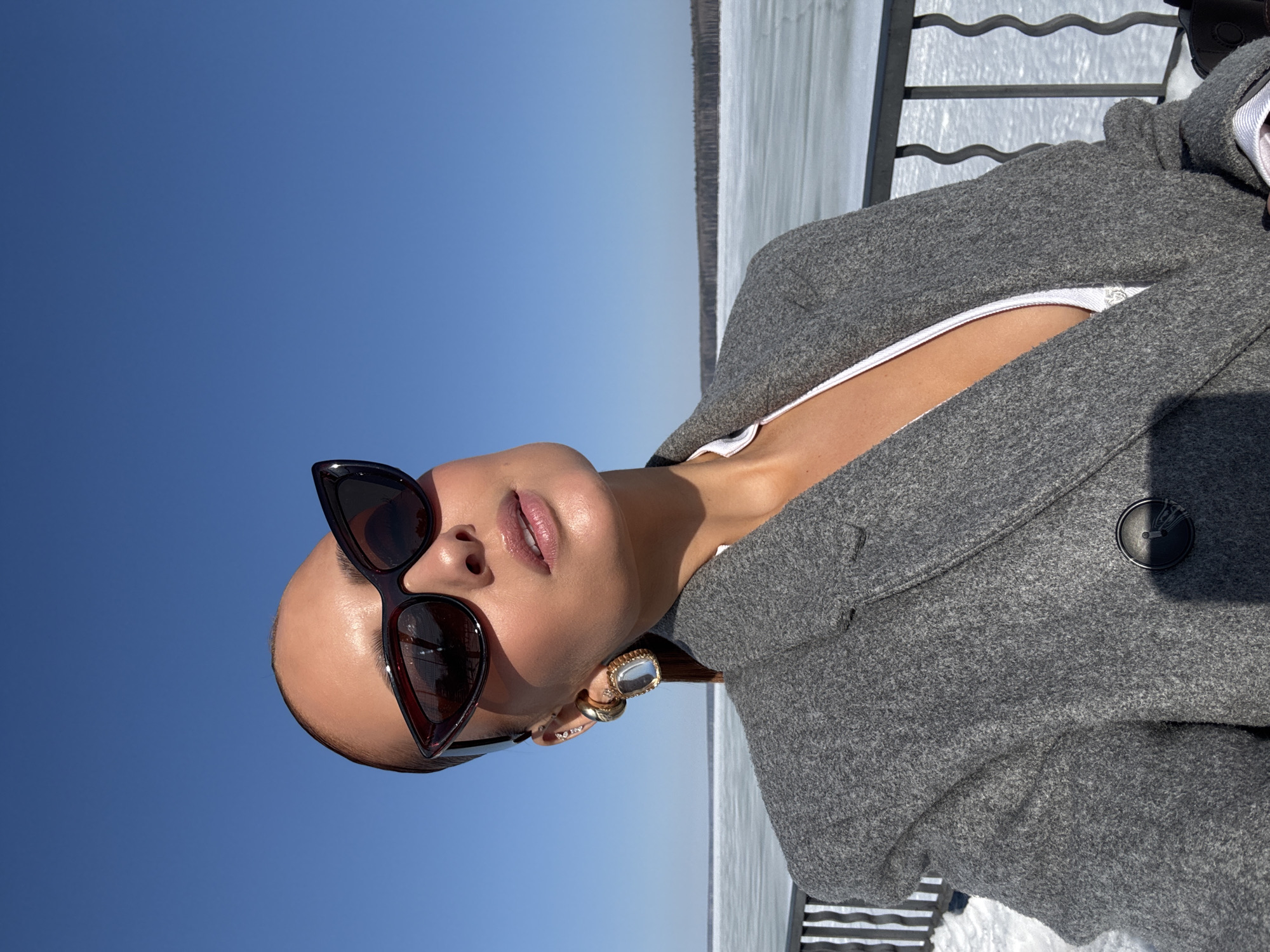 A fair-skinned female individual is captured in a close-up portrait, from the upper chest upwards, under a clear blue sky, likely at an overlook adjacent to the Votkinsk Reservoir in Chaikovsky, Russia. She is positioned with her head tilted upwards and slightly to her right, wearing dark, cat-eye style sunglasses. Her hair is pulled back. She wears a grey, textured blazer or coat over a white garment, which reveals a deep neckline. Her visible left ear is adorned with multiple earrings, including a prominent gold-toned rectangular drop earring with a clear stone, a smaller gold hoop, and several stud earrings higher on the ear. Her lips are slightly parted and appear glossy. In the background, a vast expanse of snow-covered frozen water, presumably the Votkinsk Reservoir, stretches towards a distant line of dense dark forest on the horizon. A dark metal railing with undulating vertical bars is visible in the lower left and right foreground, bordering a snow-covered area. The scene is brightly lit by the sun, indicating clear weather.