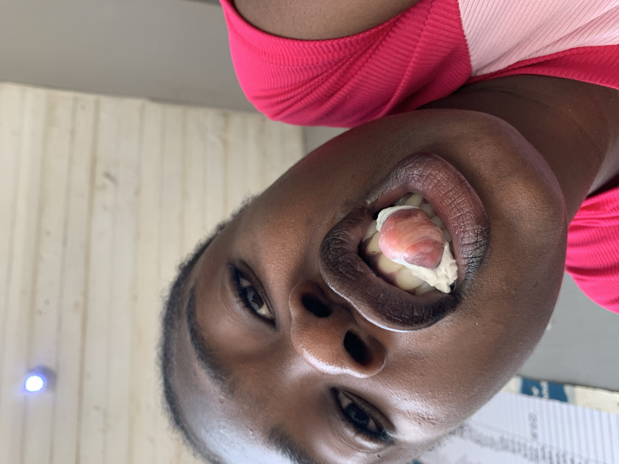 A close-up, angled shot captures a person with dark skin and dark hair, wearing a ribbed pink shirt, facing the camera at an upward angle. Their mouth is open, revealing teeth and a red and white mass, possibly related to dental work. The background is a plain wall with a light fixture visible on the left and a partially visible document or chart on the lower right. The image was taken somewhere in New Stadium, Nigeria.