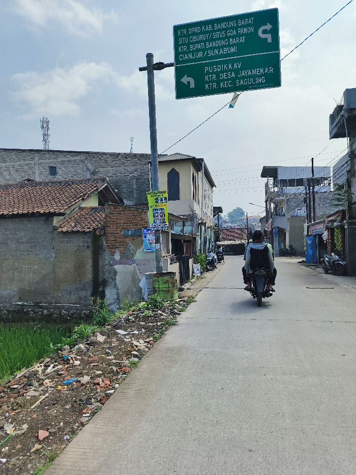 The scene is a street in Padalarang, Indonesia, located at the coordinates -6.853 latitude and 107.477 longitude. The street is paved and lined with low-rise buildings, some of which appear to be residential. There is a signpost on the left side of the street with two green signs. The top sign reads "KTR. DPRD KAB. BANDUNG BARAT SITU CIBURUY/ SITUS G.A. PANON KTR. BUPATI BANDUNG BARAT CIANJUR / SUKABUMI". The bottom sign reads "PUSDIKAV KTR. DESA JAYAMEKAR KTR. KEC. SAGULING". A man is riding a motorcycle down the street, heading towards the right side of the frame. The ground on the left side of the street is covered with a layer of dirt and debris. There is a small patch of green grass visible in the lower left corner of the frame. Several buildings line the street, some with windows and some without. The sky is partially cloudy. There is a tall thin pole with a device at the top, likely a radio antenna. There are several signs visible on the buildings. There is a trash can on the left side of the street, next to the signpost. There is another motorcycle parked on the street on the right side of the frame.