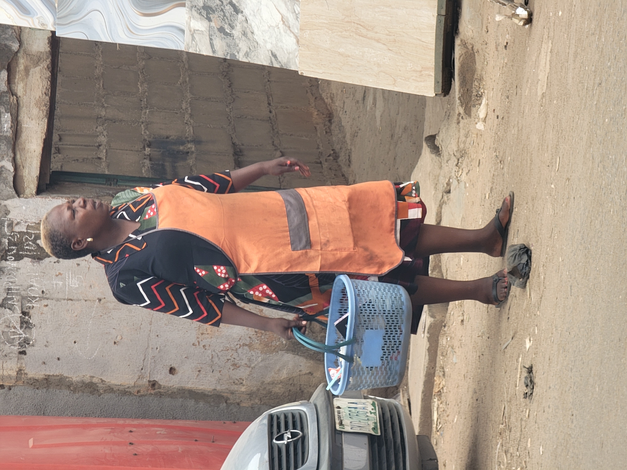 A woman wearing a black patterned shirt with an orange apron, sandals, and holding a blue plastic basket containing trash is standing on a paved street next to a gray car. The car's license plate is partially visible and appears to have an expiration date of 2024. The woman's gaze is directed to the left. A weathered, light-colored building facade with possible graffiti is in the background. This scene is taking place in Maiburiji, Nigeria.