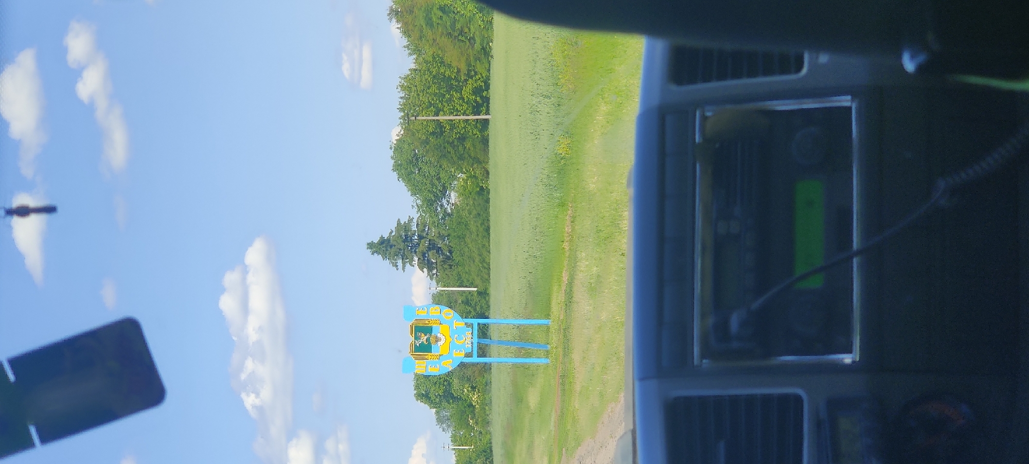 This image, taken from inside a vehicle, captures a bright, clear daytime scene. In the foreground, elements of a vehicle's dashboard are visible, including dark air vents and electronic equipment with a rectangular green light and a coiled cable.
The primary subject in the midground is a distinctive blue and yellow welcome sign. The sign is shaped like a stylized horseshoe or archway, supported by two blue posts. It features a central crest or emblem with a green background and gold/yellow elements. Prominently displayed in Cyrillic script on the sign is the text "БЕРЕЗІВ" (Bereziv). The blue and yellow colors of the sign could be indicative of Ukrainian cultural influence or location.
Behind the sign, a vast green field or meadow stretches towards the horizon. A line of lush green trees forms the background, beneath a bright blue sky dotted with scattered white, fluffy clouds. The overall setting suggests a rural or semi-rural landscape, possibly at the entrance to a town or village. The clear weather and vibrant natural colors indicate a pleasant day.