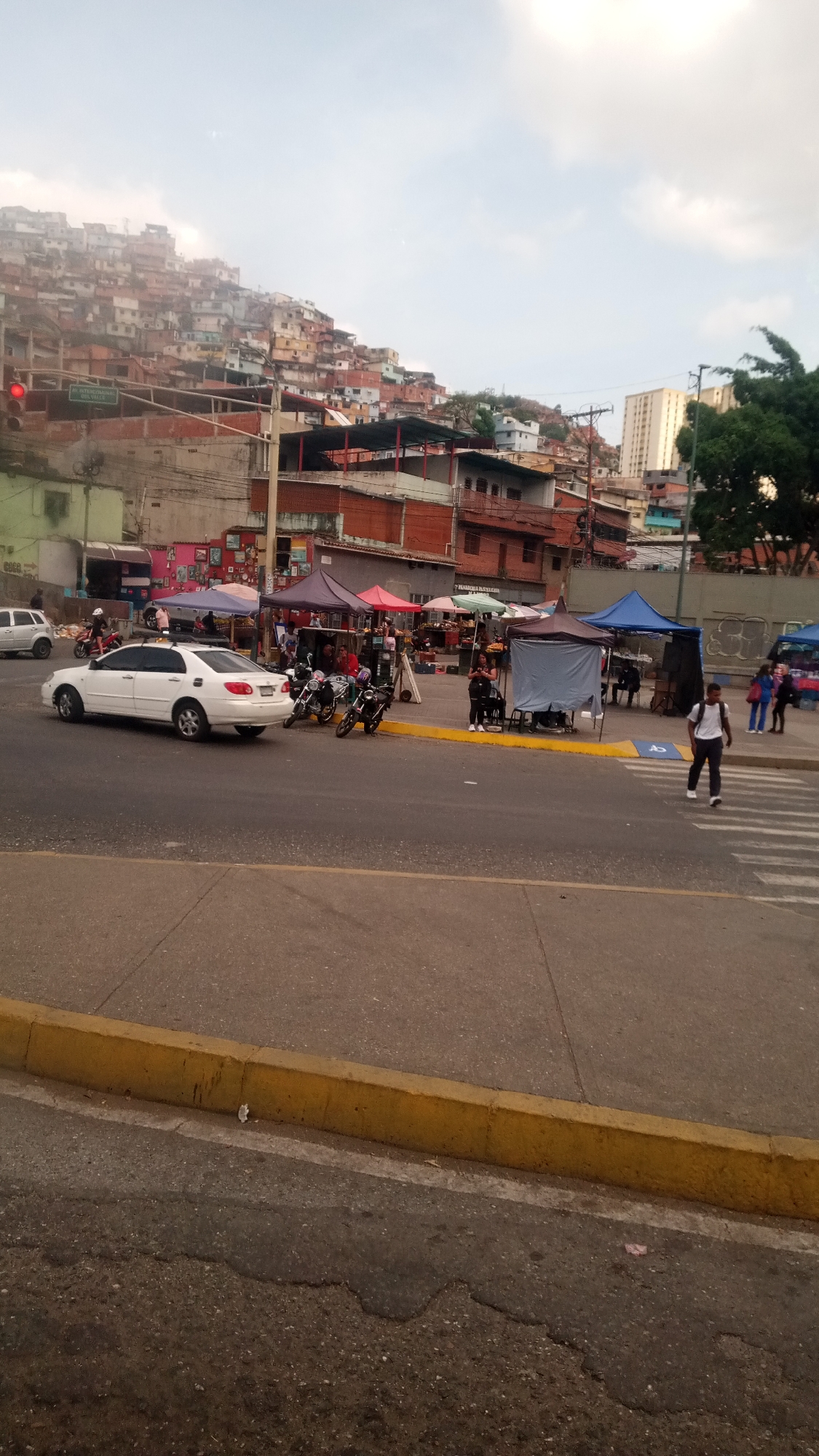 The image shows a street scene in an urban area, likely a city in Venezuela. A white car is stopped in the left lane of the road. A street market is set up on the sidewalk, with multiple vendors under colorful tents. Several motorcycles are parked near the market stalls. A man in a white shirt and dark pants is crossing the street on a crosswalk. In the background, a hillside is densely populated with buildings, characteristic of informal settlements or "ranchos." A tall building is also visible in the background. The sky is overcast.