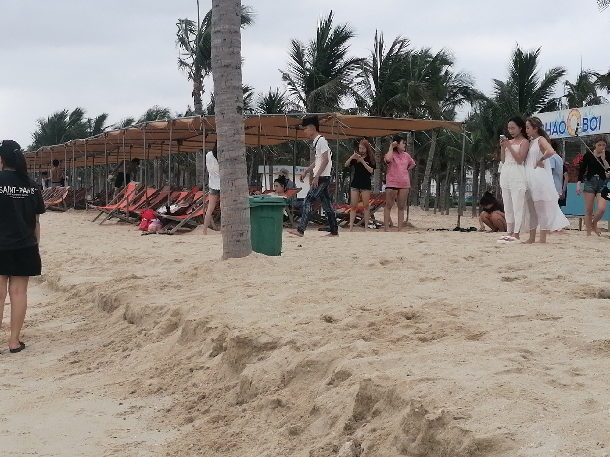 The image depicts a leisure scene on a sandy beach in Bai Chay Ward, Vietnam, under an overcast sky. The foreground features light-colored sand with minor undulations. In the mid-ground, a series of long shade structures with light-brown canopies provide shelter, beneath which several red and orange beach chairs are arranged. Numerous palm trees form a dense green backdrop behind these structures.
Many people are visible enjoying the beach. In the immediate foreground on the left, a woman in a black t-shirt and black skirt stands with her back to the camera. Her shirt displays the text "SAINT-PARIS". Further into the scene, beneath the canopies, individuals are seated in chairs or walking. A man in a white t-shirt and jeans walks past a green trash can. To the right, a group of young women is gathered; two in white attire are focused on a smartphone, while others in casual wear, including a pink top and shorts, and a black top and shorts, stand nearby or crouch on the sand.
The general activity suggests relaxation and socializing. The weather appears mild with a cloudy sky, hinting at either an early morning, late afternoon, or a generally overcast day. A notable object is a sign in the background with a blue and white lifebuoy graphic and partially visible Vietnamese text, "CHAO BOI". The overall impression is one of casual enjoyment at a tropical beach destination.