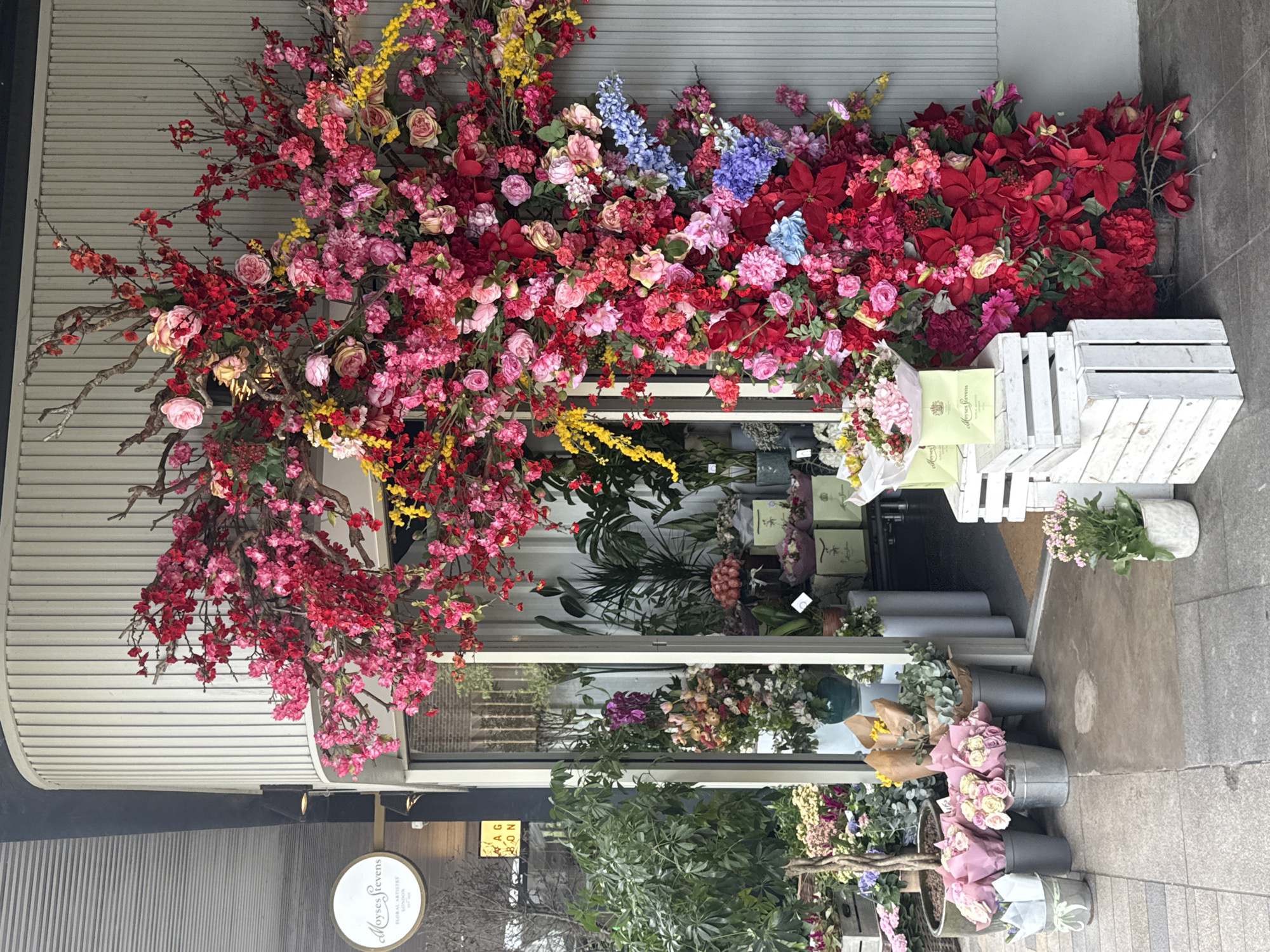 The image shows the facade of a flower shop called "Moyses Stevens" in London, United Kingdom. The shopfront is adorned with a large, colorful floral display consisting of red, pink, yellow, and blue flowers, overflowing onto the sidewalk. Several bouquets and plants are arranged in buckets and wooden crates outside the shop. The street appears to be paved with stone.