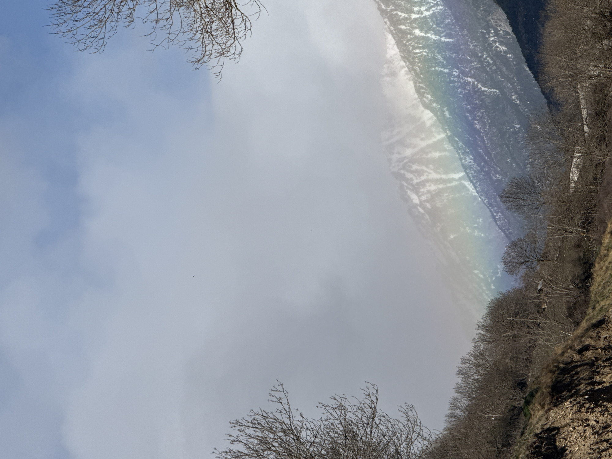 The image is a low-angle shot of a mountain scene in Sahún, Spain. A rainbow appears to the right, over the hillside. The hillside is covered in leafless trees and ground vegetation. The sky is cloudy and overcast. Bare tree branches are in the lower and upper corners of the view. Snow and a mountain are visible in the background.