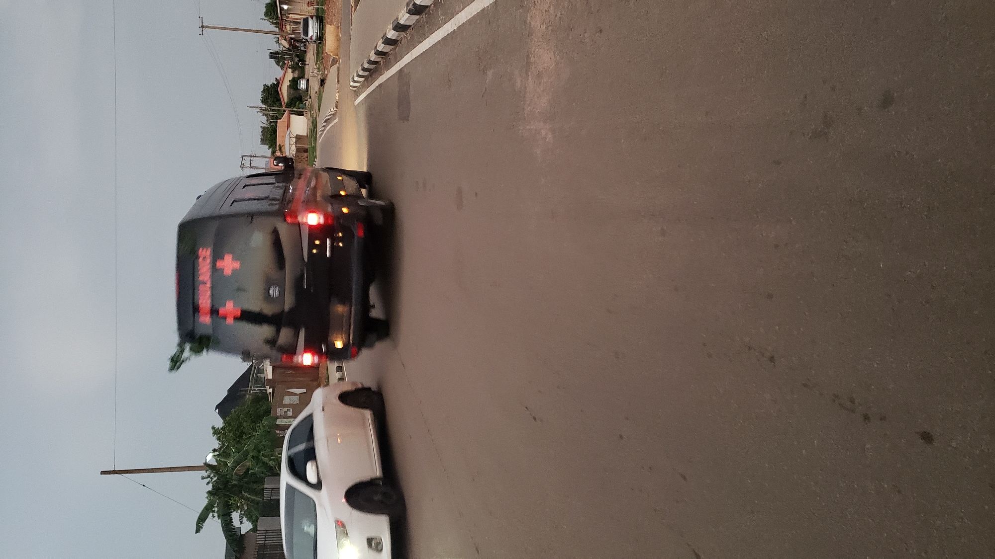 An ambulance is seen in the street of Narayi, Nigeria. The ambulance is closely followed by a white car. The road is paved, and black and white striped barriers border it. Buildings and utility poles are visible on either side of the street. The sky is overcast.
