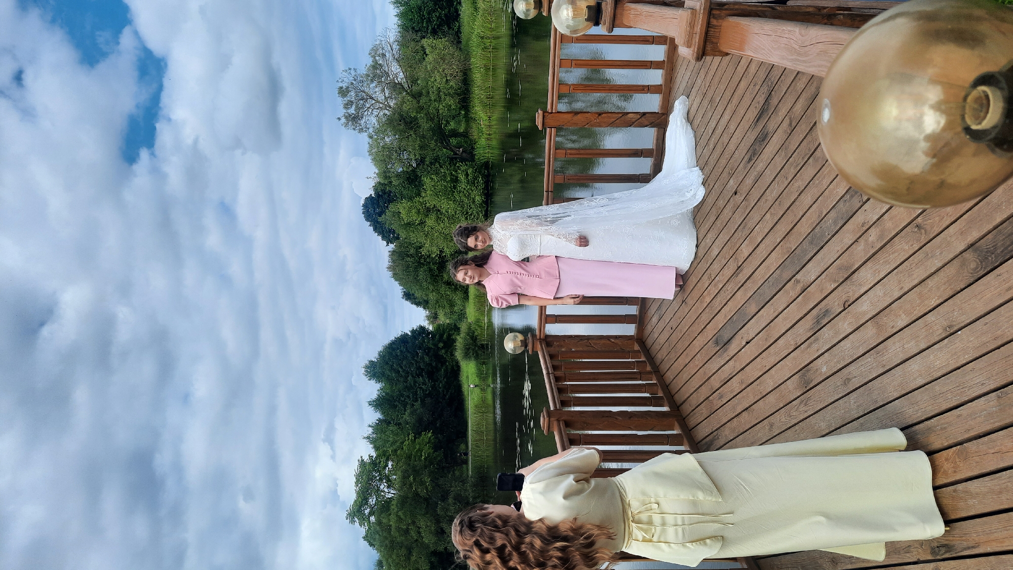 The image depicts a wooden dock with a body of water and trees in the background. The sky is overcast with clouds. On the dock, there are three individuals. In the foreground, on the left, a person with long curly brown hair is holding up a phone to take a picture. This person is wearing a long, pale yellow dress. In the middle ground, two women are posing for the photograph. One woman, on the left, is wearing a pink dress with a high collar and decorative buttons. The woman on the right is wearing a white wedding dress with intricate lace details and a long veil. The dock is made of wooden planks and has a wooden railing. There are decorative glass-covered lamps on the railing. The scene appears to be outdoors in a natural setting, likely a park or a waterfront area. The location is specified as Жодзішкаўскі сельскі Савет, Belarus.