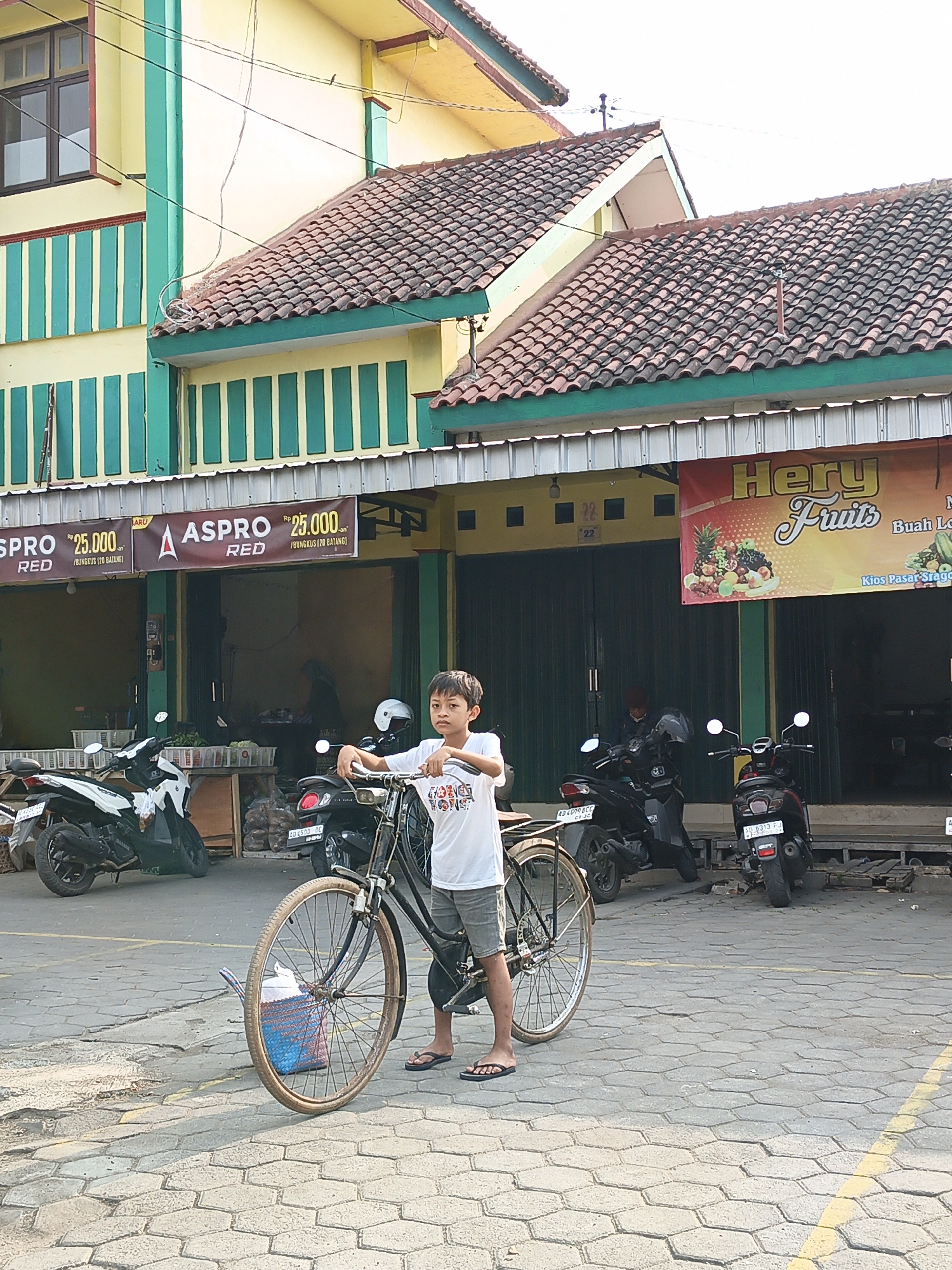 A young Indonesian boy stands next to a bicycle on a paved street in front of a building that appears to be a shop or market stall in Klaten, Indonesia. The boy is wearing a white t-shirt and gray shorts, with flip-flops on his feet. The bicycle is an older model, with a dark frame and a basket on the front. Behind the boy, the building has a yellow facade with green trim and tiled roofing. There are several motorcycles parked in front of the building, and a sign that reads "Hery Fruits" is visible. The street is paved with hexagonal stones. The overall impression is of a bustling marketplace scene.
