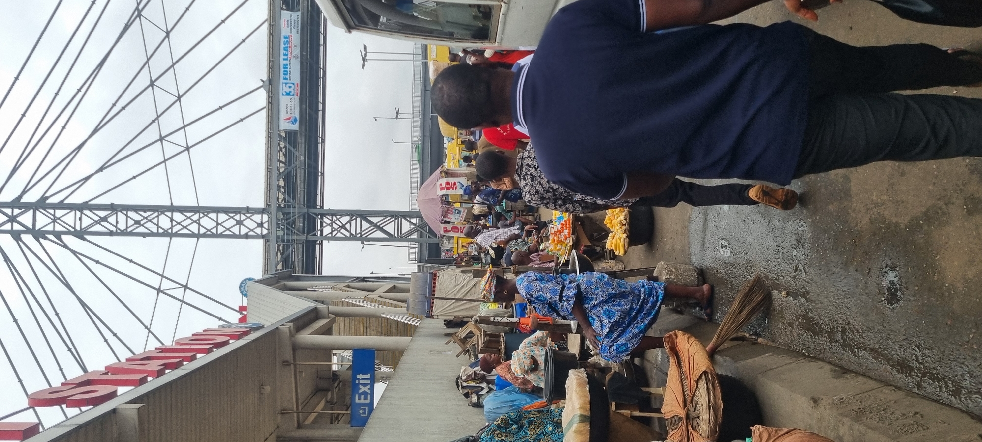 This image captures a bustling street scene in Ikeja, Nigeria, likely near a bus station as indicated by the "STATION" signage and the presence of various buses. The foreground is dominated by a busy marketplace where vendors are selling their goods. In the center, a woman in a blue and white patterned dress sits on a raised platform, observing the activity. Adjacent to her, a stall displays an abundance of corn.
To the left, an "Exit" sign points the way out of a building, suggesting the area is a transportation hub. The environment appears somewhat weathered, with a wet, possibly muddy, ground surface and various stalls and makeshift structures that create a vibrant, yet somewhat chaotic, atmosphere. People are seen walking, standing, and interacting, suggesting daily commerce and transit. The background features a large metal structure, likely part of the overhead bridge or station architecture, with a "FOR LEASE" banner visible, indicating commercial activity and development in the area. The sky is overcast, contributing to the overall dim lighting of the scene.