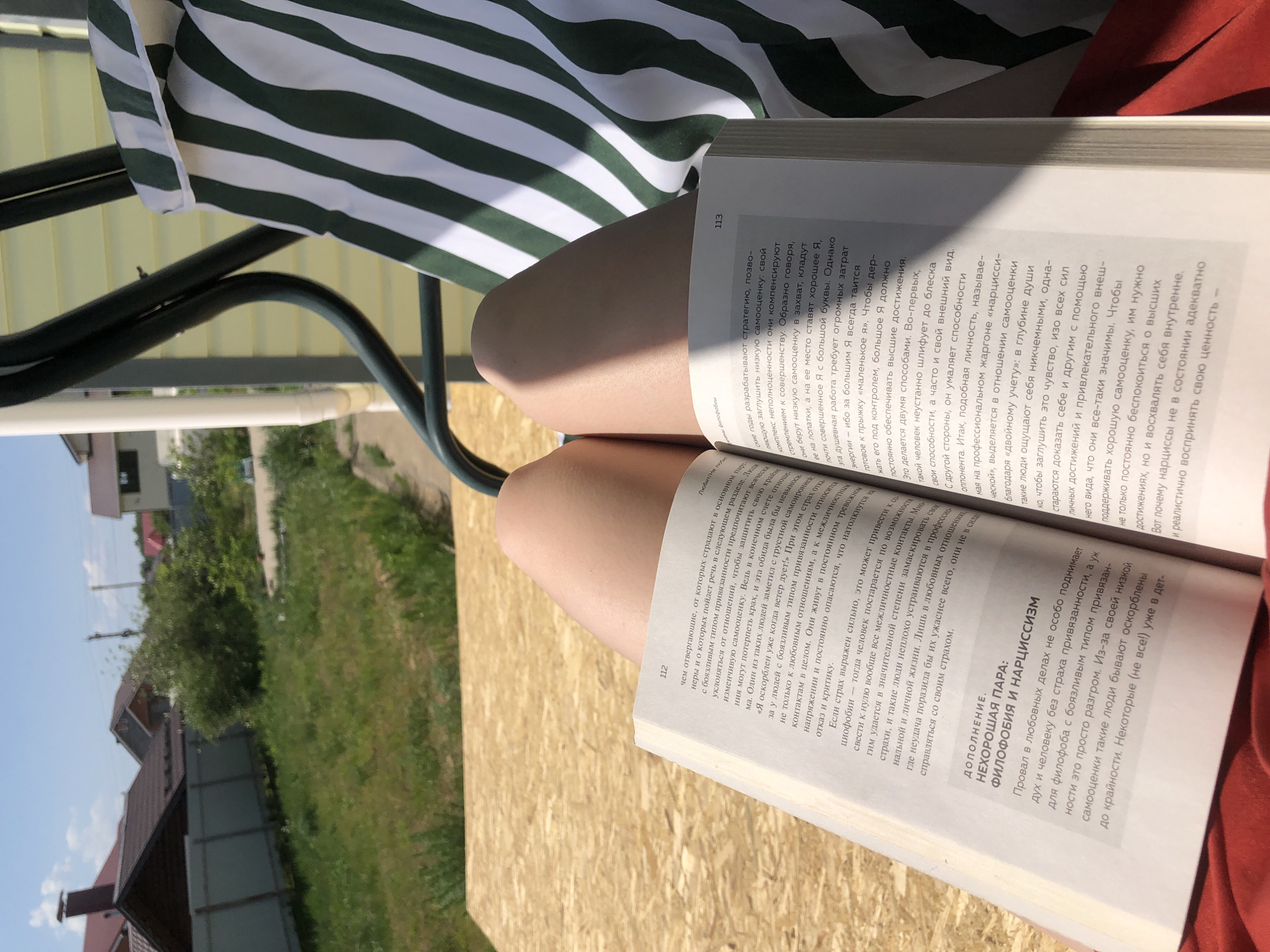 The image shows a person sitting outdoors, likely on a porch swing or similar seat, with an open book resting on their thighs. The book is in Russian, with visible text discussing psychology, specifically "philo-phobia" and "narcissism." The page on the left is numbered 112, and the one on the right is numbered 113. The person's legs are fair-skinned and bare.
Behind the person is a green and white striped awning. The background features an out-of-focus view of a residential area with green grass, trees, and rooftops, suggesting a suburban or rural setting, and potentially the city of Noviy, Russia. It is daytime and the lighting indicates it is sunny.
