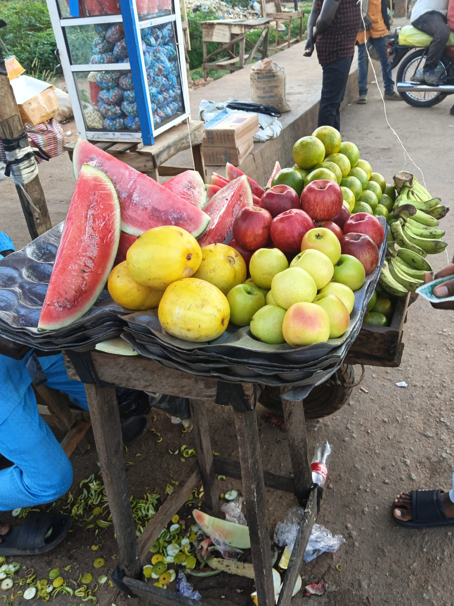 The image depicts a street market scene in Bukuru, Nigeria, showcasing a vibrant display of fresh produce. A wooden cart, laden with fruits, stands prominently. Watermelon slices, some still wrapped in plastic, are arranged on a dark mat. Alongside them are yellow mangoes, red apples, and green oranges, piled high in separate sections. Bunches of green bananas hang from the side of the cart. In the background, a glass-fronted display cabinet filled with packaged goods suggests a small shop or stall. Several individuals are present, including a person in blue trousers and black sandals to the left, and another person partially visible to the right, wearing black sandals and holding a small, light-colored object, possibly currency or a receipt. The ground around the cart is dusty and littered with discarded fruit peels and debris. The overall environment appears to be an outdoor marketplace, with a backdrop of greenery and other stalls or structures suggestive of a bustling urban setting.