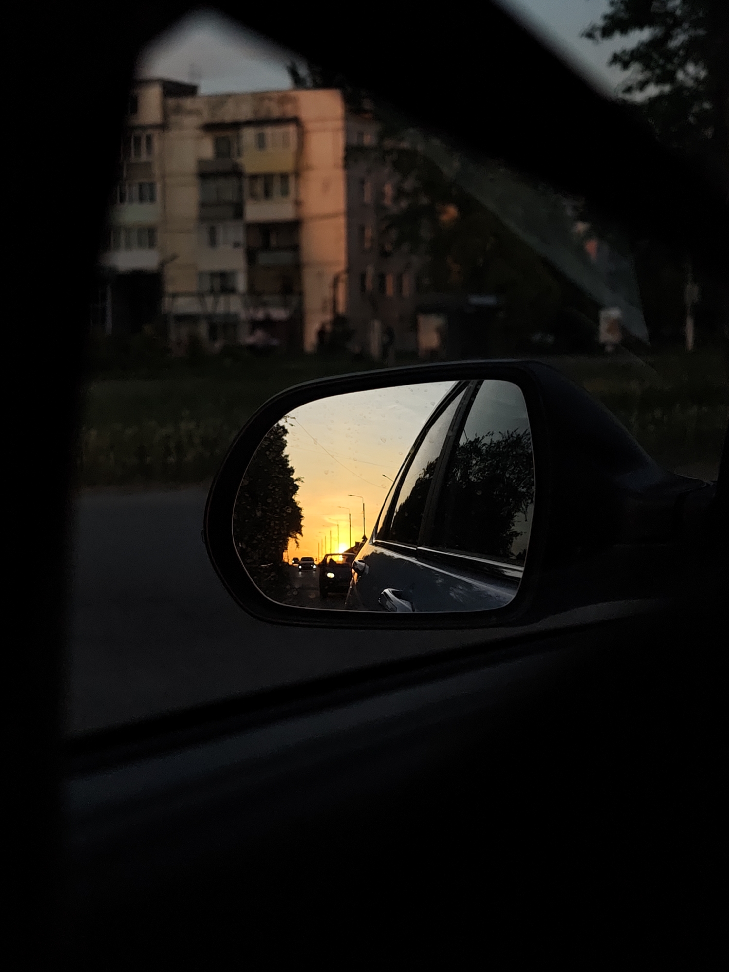 The image is a view from inside a car, looking out the side window and through the side mirror. The mirror reflects a road receding into the distance, with a bright sunset at the end of the road. Cars are visible in the mirror, driving on the road. The buildings in the reflection, as well as the buildings visible through the side window, suggest the location is in a densely populated urban area, possibly Chaikovsky, Russia. A multi-story apartment building is visible through the car's side window. The environment appears to be late evening or early morning, indicated by the low light and sunset. There are trees alongside the road. The interior of the car is dark, with the side mirror and window frame prominent in the composition.