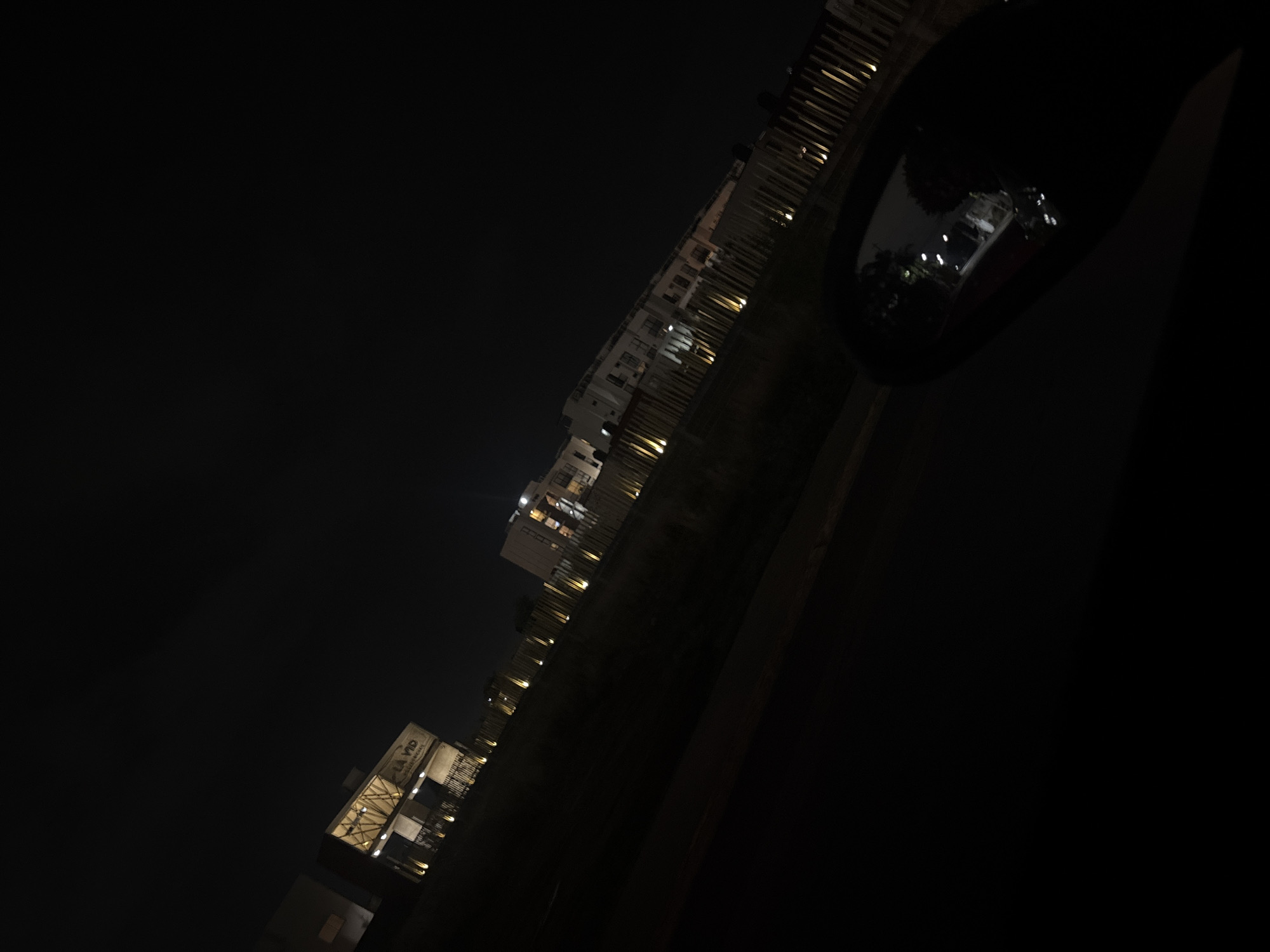The image captures a nighttime scene, viewed from inside a vehicle, possibly from a passenger's seat looking out the side window. The perspective is at a steep diagonal angle, with the horizon line slanting upwards from left to right.
In the mid-ground, a series of multi-story buildings line what appears to be a street or pathway. Many windows in these buildings are illuminated with warm light, suggesting interior activity. The architecture appears modern, with visible balconies or external structures on some buildings. Further down the line of buildings, on the left side of the frame, one structure prominently displays visible text. The text clearly reads "LA VID RESTAURANT", indicating a commercial establishment.
The foreground is dominated by a dark strip, likely a road or ground, with faint vertical elements that could be small trees or decorative lighting poles extending along its edge. On the far right side of the image, a large, dark, and reflective surface is visible, which is unmistakably a car's side mirror. This mirror reflects a distorted view of the external environment, including some bright lights and blurred shapes, further confirming the perspective from within a vehicle.
The overall scene is very dark, indicative of a clear night. There are no visible people or specific activities other than the implied presence within the lit buildings. The setting appears to be an urban or suburban area in San Francisco Ocotlán (Ocotlán), Mexico.