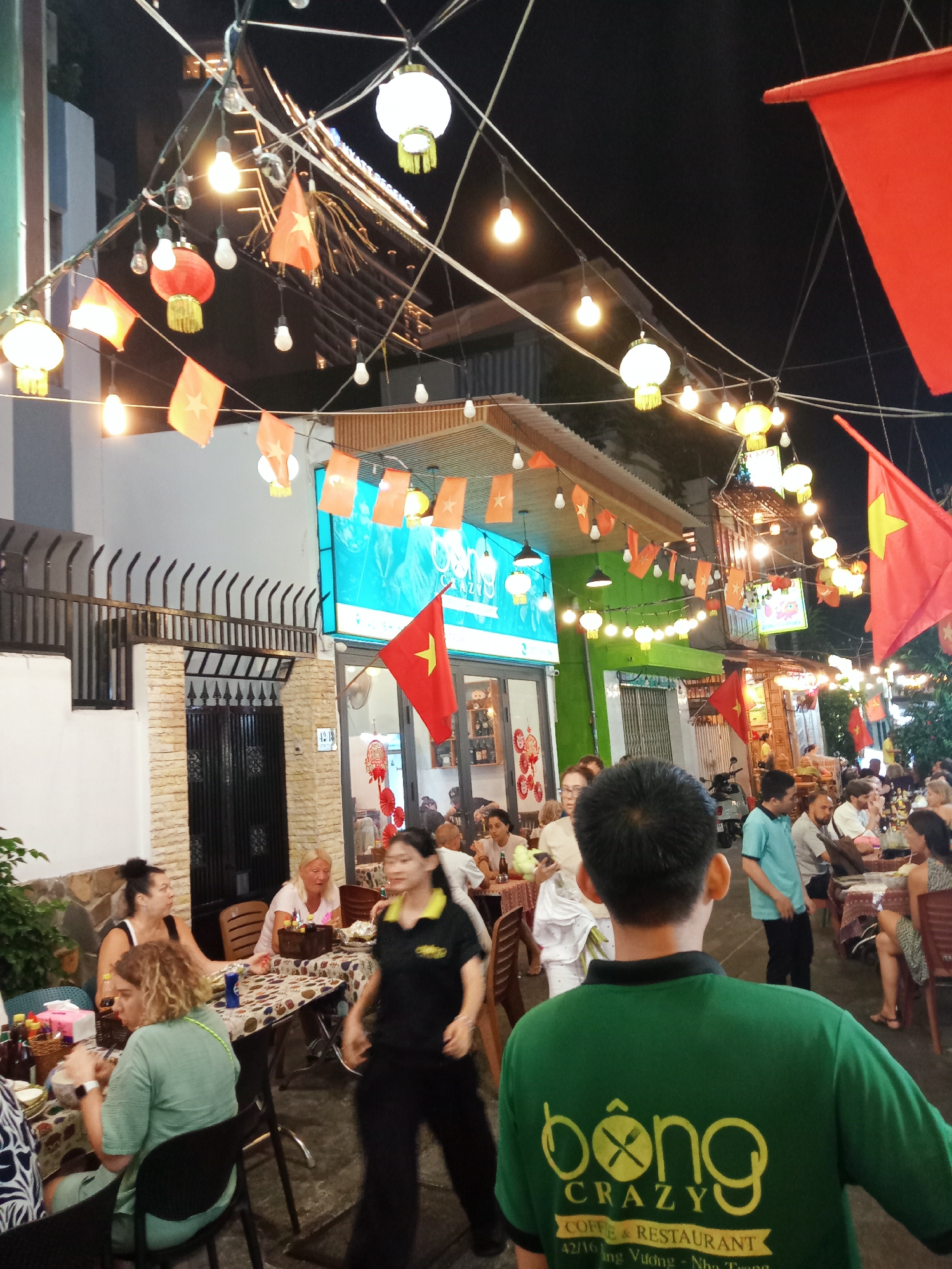 The image captures a street scene in Nha Trang Ward, Vietnam, at night. The street is lined with open-air restaurants and is heavily decorated with Vietnamese flags, string lights, and lanterns. People are seated at tables, dining and interacting. A waiter in a green shirt with "bông CRAZY" logo on the back walks through the scene. The restaurants are on either side of the street, and buildings are painted in white, green and other light colors.