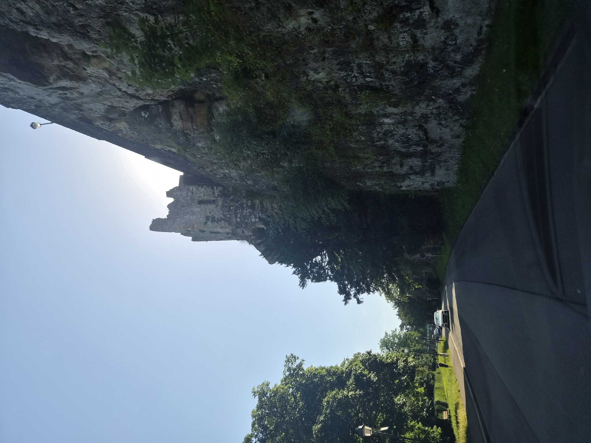 A road curves along the base of a large, weathered stone wall, partially overgrown with vegetation, in Luxembourg City, Luxembourg. The wall appears to be part of historical fortifications, with a section of ruined structure visible at the top. A lamppost is mounted on the wall's edge, and sunlight streams from behind the wall, creating a lens flare effect. On the other side of the road, a row of lush green trees lines a grassy area with a sidewalk and a park bench. Several cars are visible on the road, and a few pedestrians are walking on the sidewalk. The sky is clear and blue.
