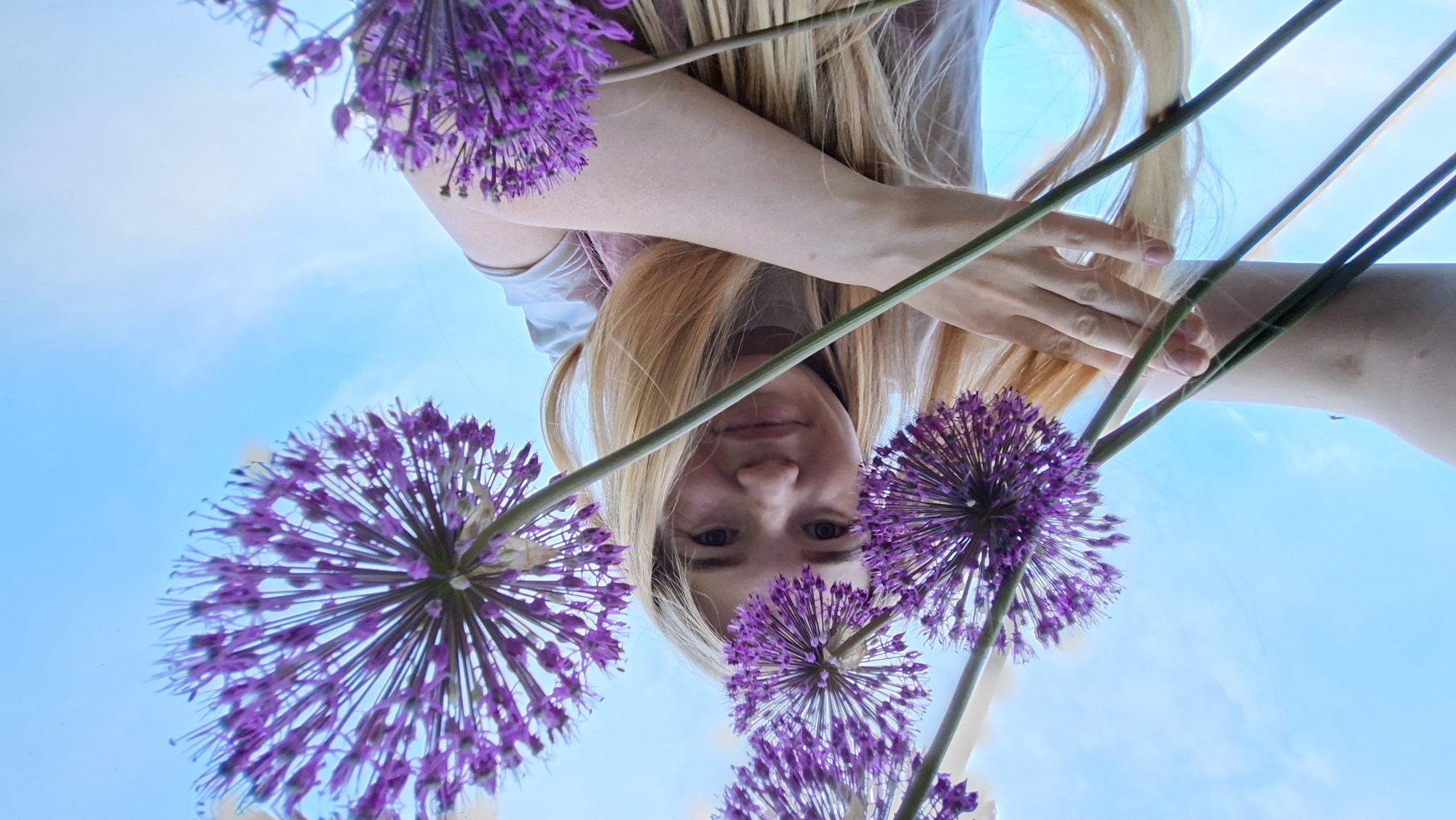 A low-angle shot depicts a fair-skinned person with blonde hair holding purple allium flowers against a blue sky. The person's face, tilted downward, is centered in the frame. The flowers, with their spherical heads, are positioned around the person's face. The sky is visible in the background, with a few clouds. No specific location details related to Большой Букор, Russia, are identifiable.