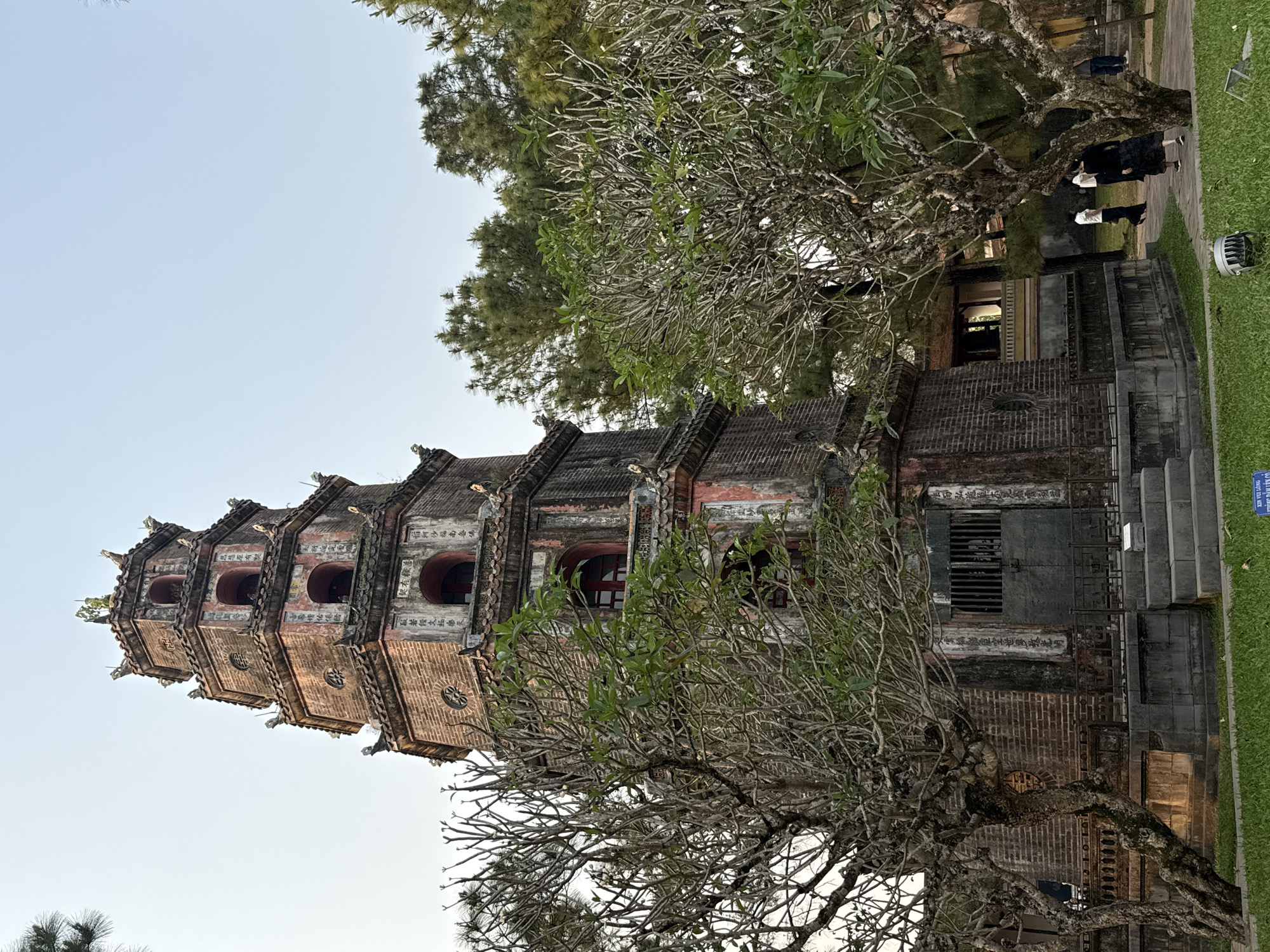 The image captures the iconic Thien Mu Pagoda, located in Kim Long Ward, Thua Thien Hue, Vietnam. The pagoda, constructed with brown brick, rises prominently against a pale sky. It is composed of tiered segments, each detailed with architectural ornaments and inscriptions, and a small arched window in each tier. A bare tree with sparse foliage partially obscures the pagoda's base, framing the structure with its branches. To the right, a glimpse of a grassy area and a pathway occupied by several people can be observed. The scene is illuminated by soft, natural light, emphasizing the ancient architecture.