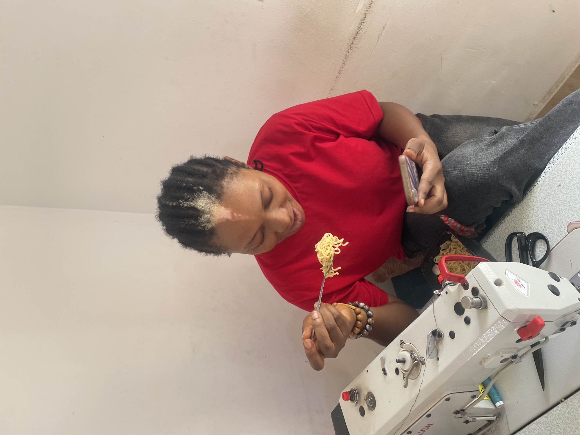 An overhead, slightly rotated shot captures a person, likely a woman, with braided dark hair that includes some light-colored sections, wearing a red t-shirt and dark grey trousers. She is seated and actively eating, holding a fork with a mouthful of noodles suspended near her mouth. In her left hand, she holds a smartphone. Bracelets made of wooden and pearl-like beads are visible on her right wrist.
To her right, on a work surface, sits a bowl of noodles and a pair of black scissors. A prominent white industrial sewing machine, featuring red accents and black controls, occupies the lower right portion of the frame. Visible text on the machine includes "PRO TION" and "LION", likely part of a brand name. Spools of blue and yellow thread are seen near the base of the machine. The setting appears to be an indoor workshop or a room dedicated to sewing, indicated by the presence of the industrial sewing machine. The walls in the background are plain and light-colored. The individual appears focused on her meal, suggesting a break from work. The scene takes place in Dushepe, Nigeria.