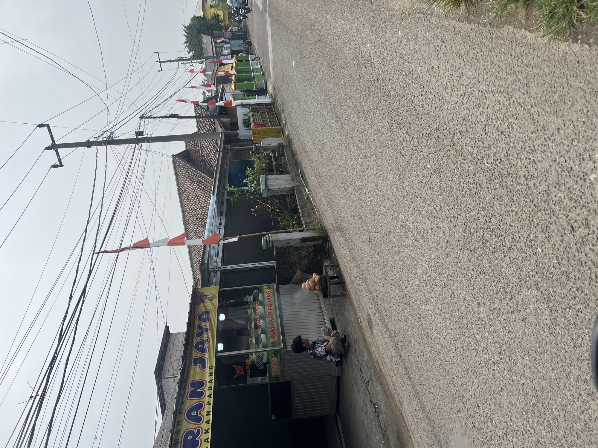 The image depicts a street scene in Klapanunggal, Indonesia, likely on an overcast day. In the foreground, a paved road runs diagonally from the lower left to the upper right. Along the left side of the road, there is a roadside food stall with a yellow awning that reads "RAN JAYA" and "MASAKAN PADANG". In front of the stall, a young man is crouched down, smoking a cigarette, and looking towards a small table on which a dish of what appears to be fried chicken is steaming. The stall itself has glass windows displaying food items. Further down the street on the right, a row of houses with terracotta roofs can be seen. Several Indonesian flags are displayed on poles along the side of the road, suggesting a national holiday or celebration. Utility poles with a dense network of electrical wires are prominent in the background. A few motorcycles and a small blue vehicle are visible further down the street, indicating ongoing traffic. The environment appears to be a semi-urban or suburban area with a mix of commercial and residential structures.