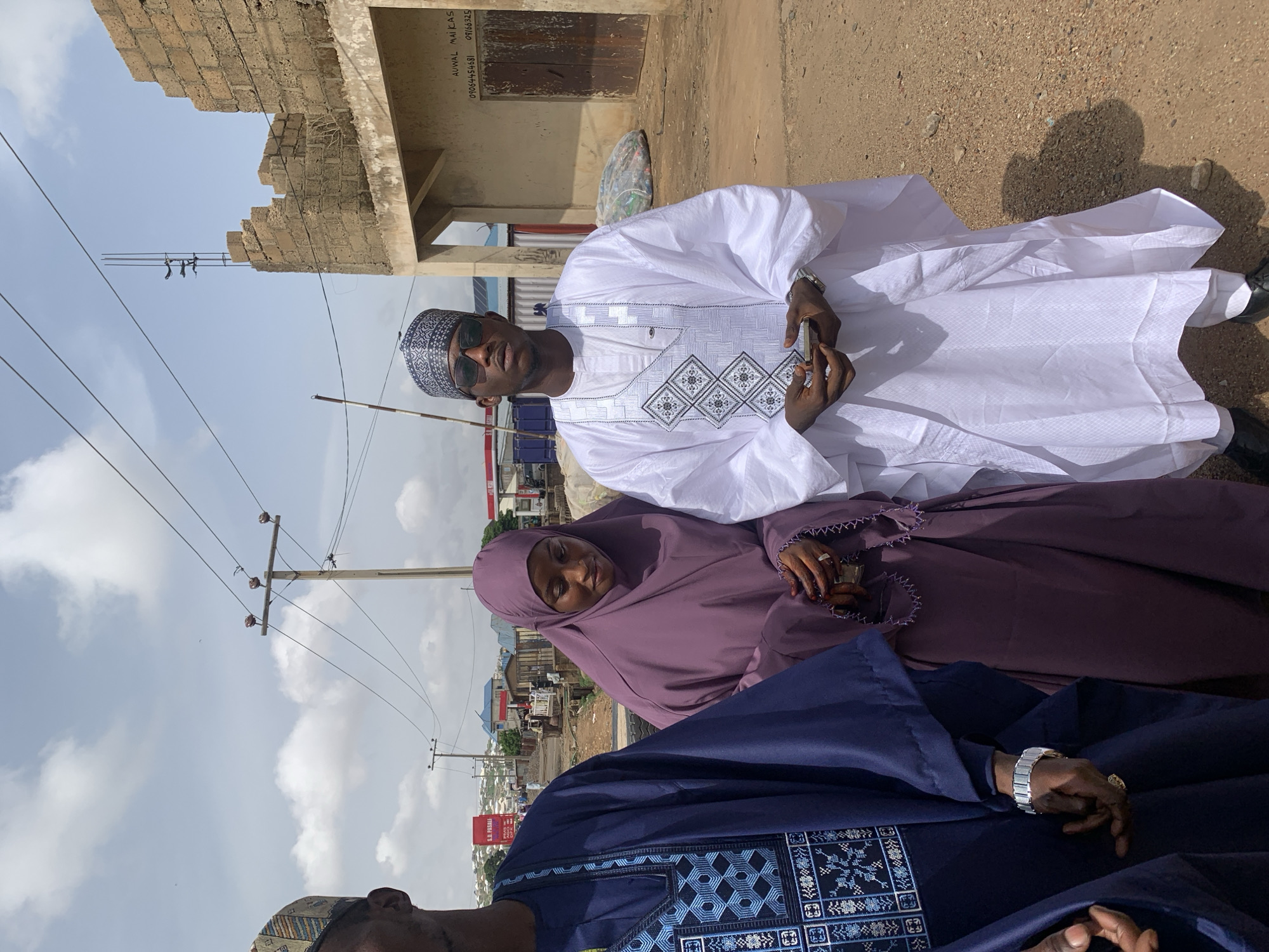 The image shows three people outside in Naraguta Hausawa, Nigeria. A man in a white, intricately embroidered traditional garment and sunglasses is holding a mobile phone. Next to him is a woman in a light purple hijab and dress, holding a small cup or glass. Partially visible at the bottom is a man in a dark blue robe with blue embroidery. The background includes a partially constructed brick building, utility poles with wires, a street, and buildings in the distance. The ground appears to be dirt or gravel. The sky is light blue with some clouds.