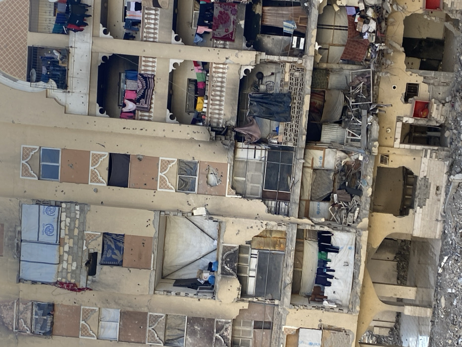 This image depicts a multi-story residential building in Khan Yunis, Palestinian Territory, exhibiting widespread structural damage. The light-colored facade is heavily marked with numerous bullet holes and impact craters across all visible floors. Extensive sections of the building show partial collapse, particularly on the lower and central-left portions, revealing exposed interiors. Many windows are either shattered or missing entirely, with several openings covered by makeshift materials including white tarpaulins, blue plastic sheeting, and fabric, some bearing the UNICEF logo. Balconies on the right side of the structure feature white balustrades and display various items of laundered clothing hanging on lines. Inside exposed rooms and on balconies, visible objects include pieces of furniture, domestic items, and general debris. The ground level directly in front of the building is covered in a dense layer of rubble, fragmented building materials, and areas of standing water. No individuals are visible within the frame.