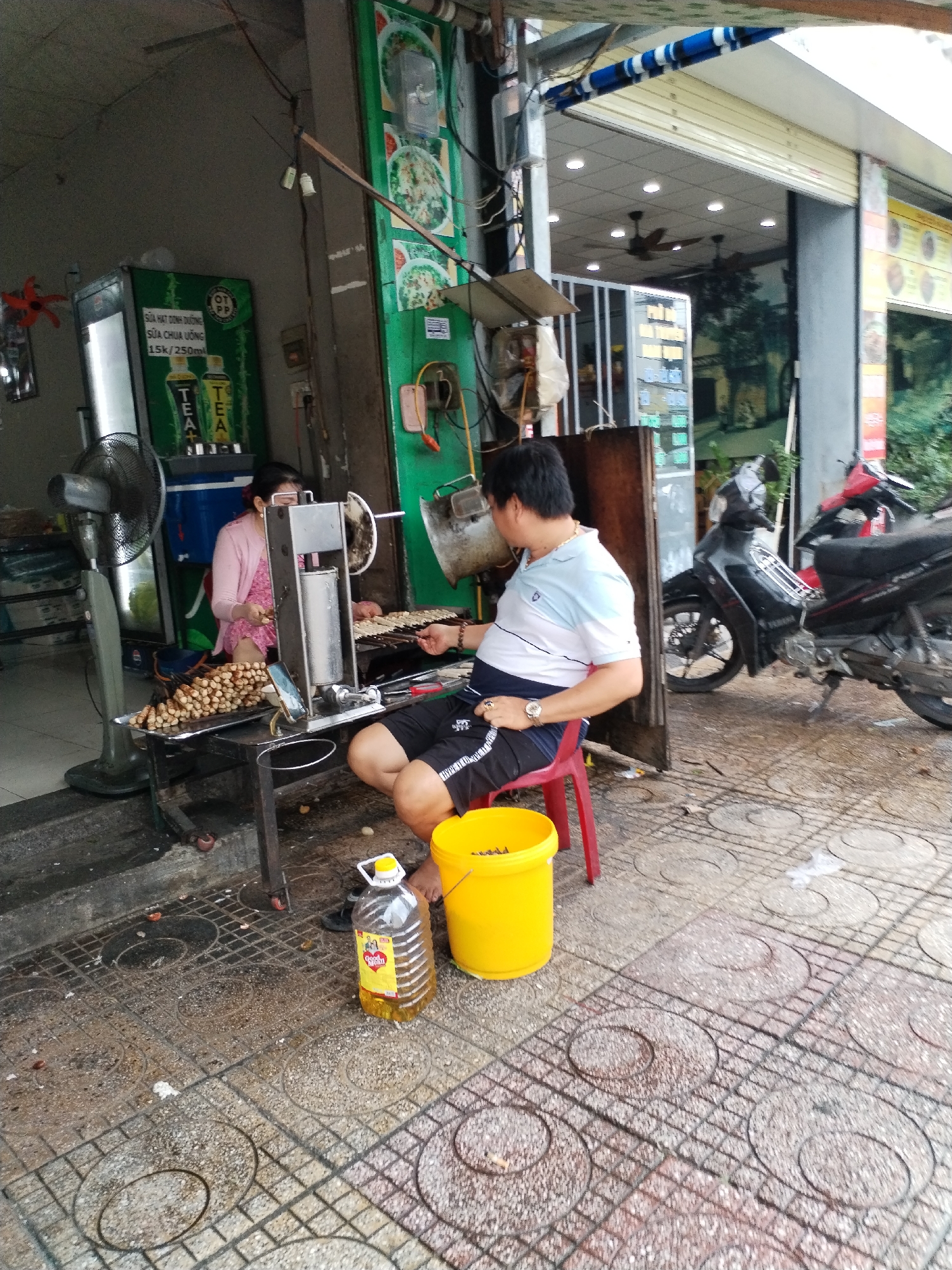 This daytime image depicts a vibrant street food scene in Nha Trang Ward, Vietnam, showcasing two individuals preparing food at an open-air stall. A woman, wearing a pink cardigan and glasses, is seated behind a metal food preparation machine, possibly used for shaping or cooking skewered items. A tray in front of her holds numerous skewers, some appearing uncooked and others seemingly grilled or roasted. An industrial fan operates beside her.
A man, wearing a white and blue polo shirt, dark shorts, a watch, and a gold chain, is seated on a red plastic stool, observing or assisting with the food preparation. His hand rests near the tray of skewers. On the patterned sidewalk tiles, a yellow plastic bucket and a large bottle of "GOLD" cooking oil are visible.
In the background, a commercial refrigerator cooler displays various beverages, including "SỮA HẠT DINH DƯỠNG" (Nutritional Seed Milk) and "SỮA CHUA UỐNG" (Drinking Yogurt) priced at "15k/250ml," along with "TEA" and "TEA+" branded drinks. Adjacent to the stall, two motorbikes are parked, and the interior of a nearby establishment with rolled-up shutters and ceiling fans is partially visible. The bright, natural lighting indicates clear, daytime weather, and the overall scene conveys the bustling atmosphere of a typical Vietnamese street food vendor.