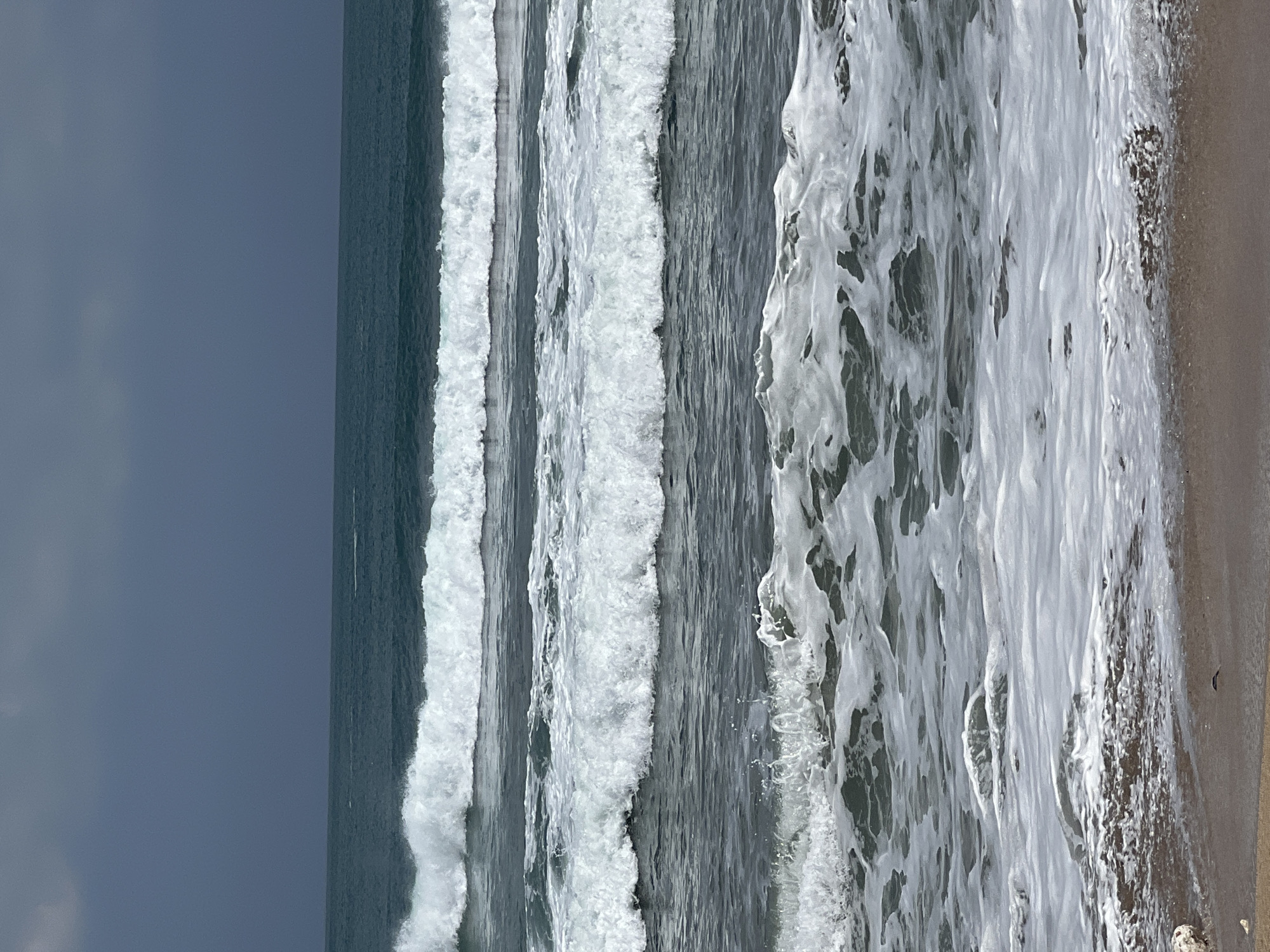 A wide shot captures multiple waves rolling onto a sandy beach in Kuta, Indonesia. The ocean is a dark teal color, contrasting with the foamy white crests of the waves. The sky above is a deep, overcast gray, with no visible sun. The waves are of varying sizes, with the closest waves being the largest and breaking with significant foam. The beach itself is a light brown sand, with some wet patches from the receding waves. There are no people or other discernible objects visible in the scene.