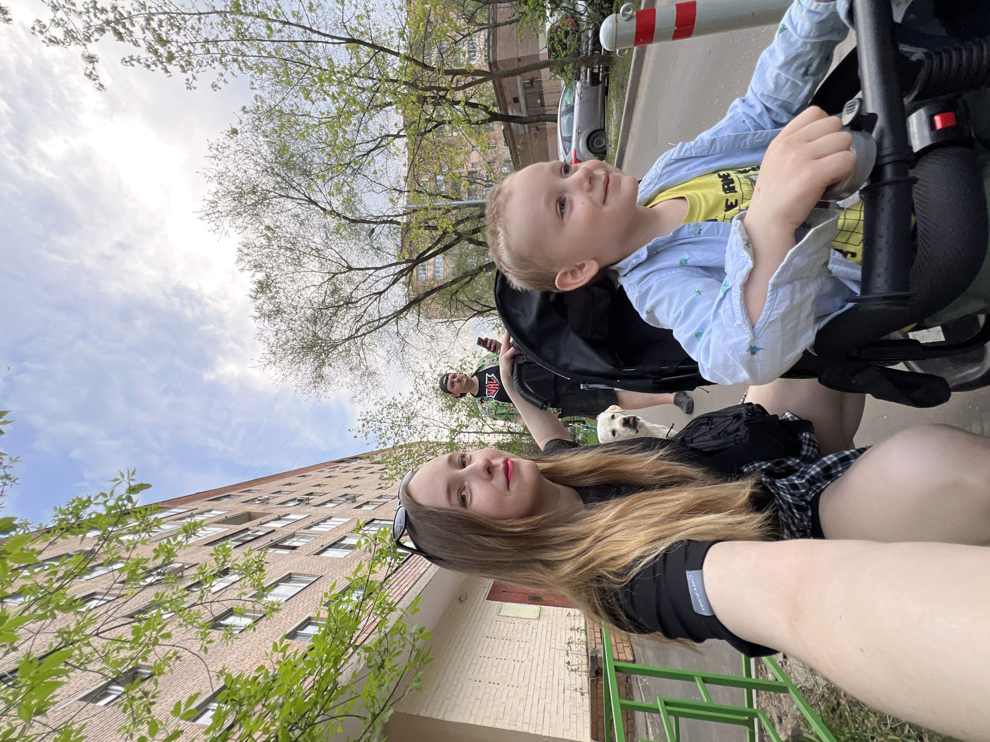 The image shows a woman taking a selfie while pushing a stroller with a young boy inside. The boy is wearing a yellow t-shirt and a light blue shirt. In the background, a man is holding a dog on a leash. The setting appears to be a residential street in Mytishchi, Russia. A multi-story brick building is visible, along with trees and a parked car. The weather is overcast with glimpses of blue sky.