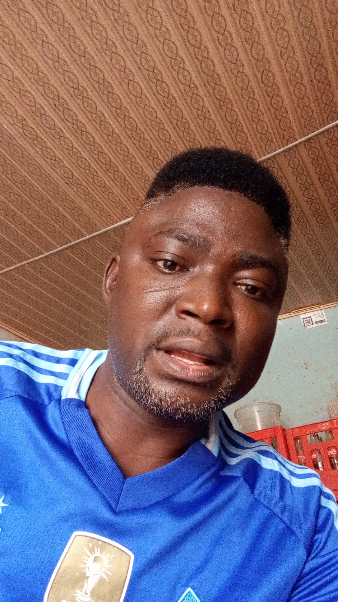 A medium close-up shot captures a man looking directly at the camera. He appears to be of African descent, with dark skin, short curly black hair showing some graying at the temples, and a short, salt-and-pepper beard. His mouth is slightly open as if he is speaking or about to speak. He is wearing a bright blue sports jersey with light blue stripes on the shoulders and sleeves. A gold emblem, prominently featuring "FIFA" and a depiction of the World Cup trophy, is visible on the left side of his chest.
The background includes a brown ceiling with a repeated, ornate pattern of interlocking loops, possibly wallpaper or paneling. A thin white cable or wire runs horizontally across the upper part of the background. To the man's right, a light blue-green wall is visible, and below it, part of a red plastic crate, possibly for beverages, can be seen. A small white label with a QR code and some indistinct text is affixed to the blue-green wall. The lighting appears to be indoor, casting subtle shadows on his face.