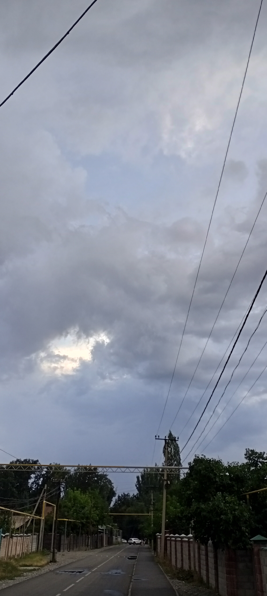 The image depicts a residential street in Sokuluk, Kyrgyzstan, under an overcast sky. Power lines crisscross the upper portion of the frame, converging on a utility pole visible in the distance. The street is paved, with some visible puddles and markings, and lined on both sides by fences and trees. Several white cars are parked or driving on the street. The overall impression is one of a quiet, possibly suburban, environment.