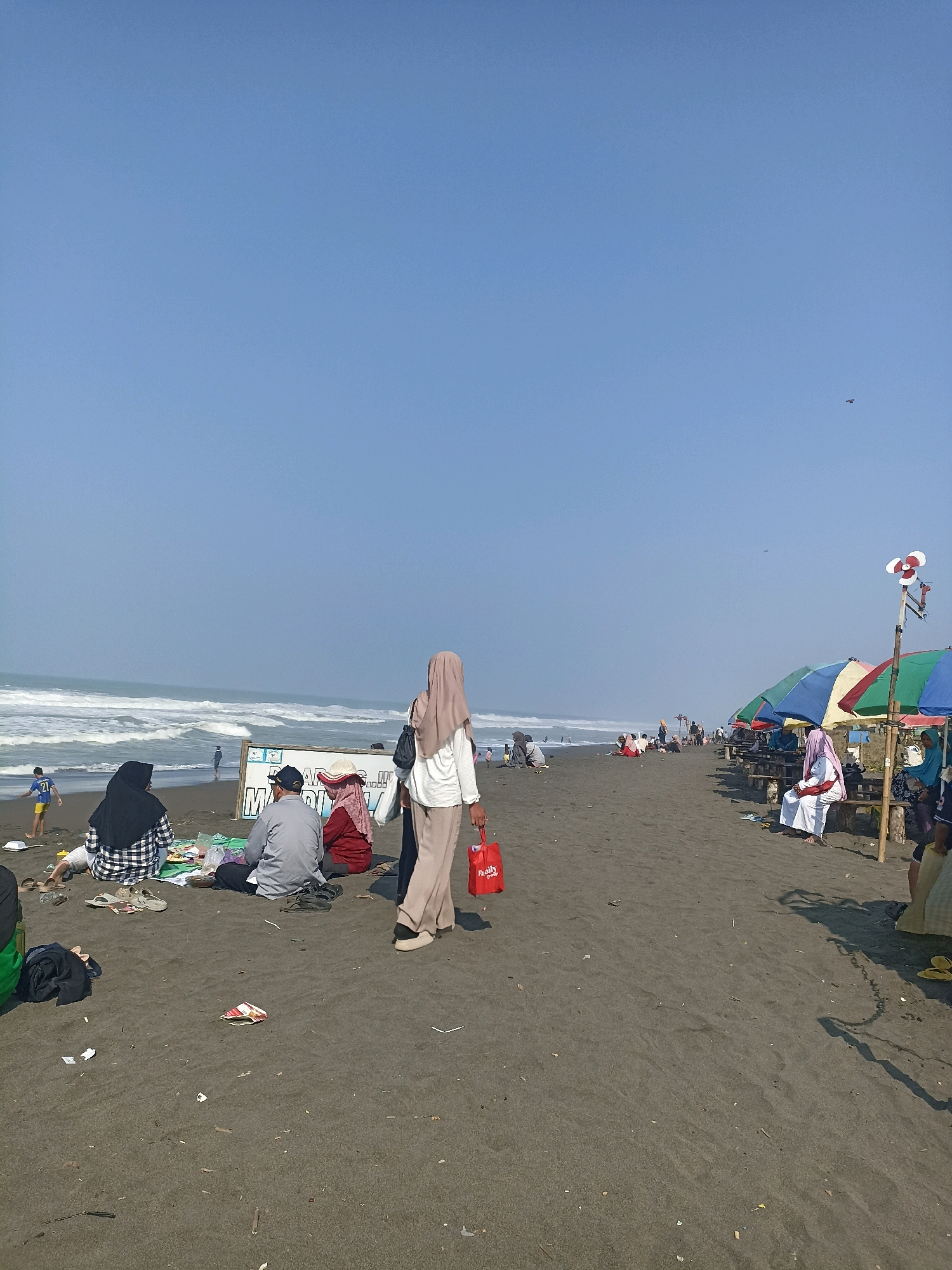 This image depicts a sunny day at a black sand beach, likely in Kenoyajaya, Indonesia. The sky is clear and blue with a few distant birds. The ocean in the background has moderate waves breaking on the shore. The beach is populated with numerous people enjoying various activities. In the foreground, a group of individuals are gathered on a mat, engaged in what appears to be a picnic. One person is wearing a black and white checkered outfit, another a grey shirt and a black cap, and a third a red shirt and a pink headscarf. Further down the beach, several colorful umbrellas are set up over wooden tables and benches, providing shade for people resting or eating. Some individuals are seated at these tables, while others are standing or walking along the sand. To the left of the image, a young boy in a blue shirt with the number 21 is playing near the water's edge. A sign with text in Indonesian is visible behind the picnic group. The overall scene conveys a relaxed and recreational atmosphere on a typical day at the beach.