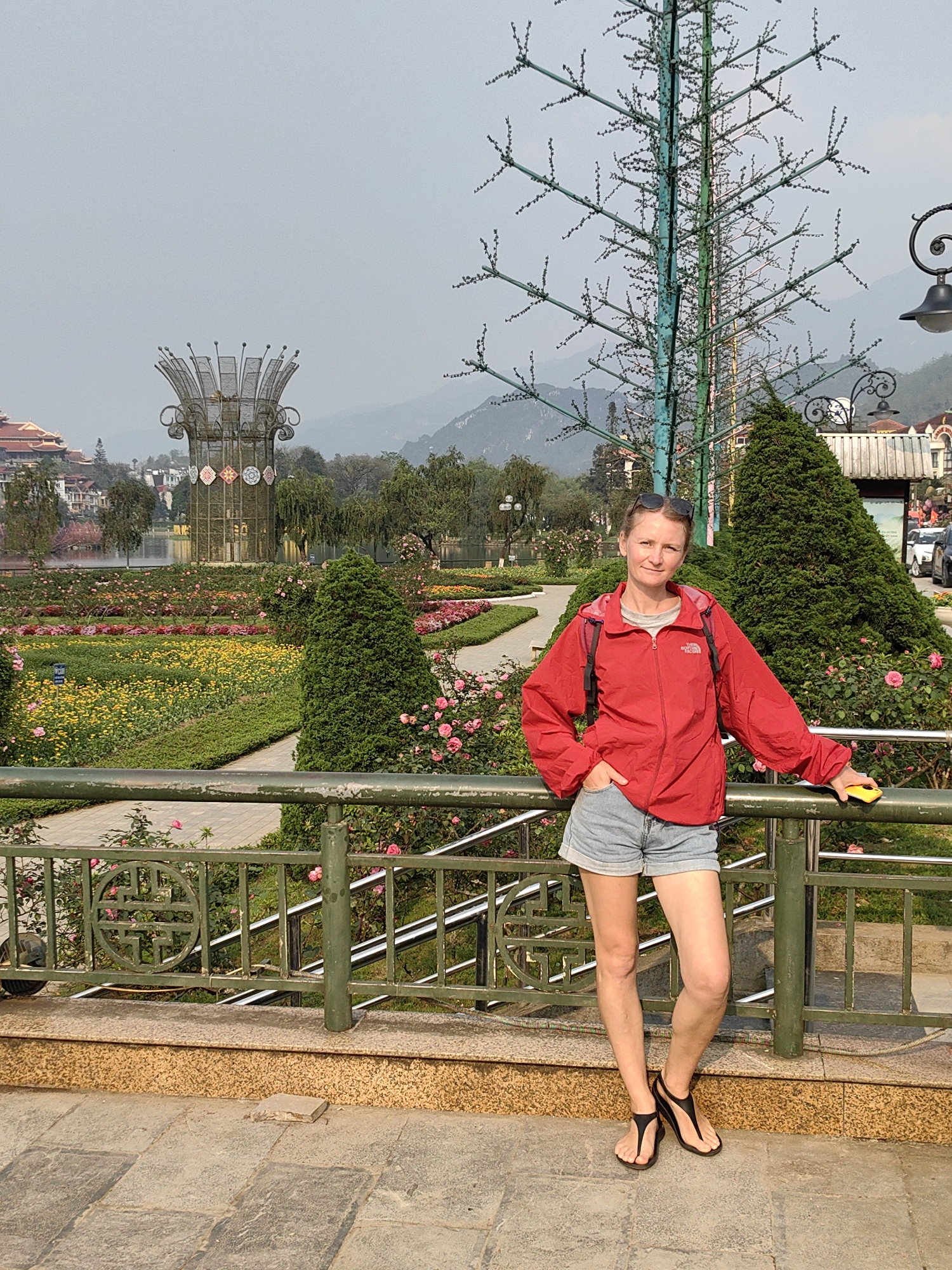 A female individual stands posing in the Sa Pa Lake Park and Flower Gardens in Sa Pa, Vietnam. She is positioned behind a dark green metal railing with ornamental circular motifs, resting her right arm on it while holding a yellow smartphone. Her left hand is in her pocket. She wears a red full-zip jacket with a visible "The North Face" logo, a grey t-shirt, light blue denim shorts, and black thong-style sandals. Sunglasses are pushed onto the top of her head.
Behind the individual and the railing, a meticulously maintained public garden unfolds, featuring paved walkways, conical evergreen shrubs, and flower beds densely planted with yellow flowers and pink roses. In the mid-ground, a tall, stylized metal tree structure, painted green and blue with small decorative elements, stands to the right. A prominent, large metal sculpture resembling a crown or stylized lotus, adorned with patterned panels at its base, is situated further back in the garden, adjacent to Sa Pa Lake (Hồ Sa Pa). Traditional-style buildings with multi-tiered red roofs are visible on the far side of the lake, against a backdrop of hazy, distant mountains under a light grey sky. Vehicles are partially visible on a road in the far right background. The foreground consists of grey stone paving and a textured golden-brown curb.