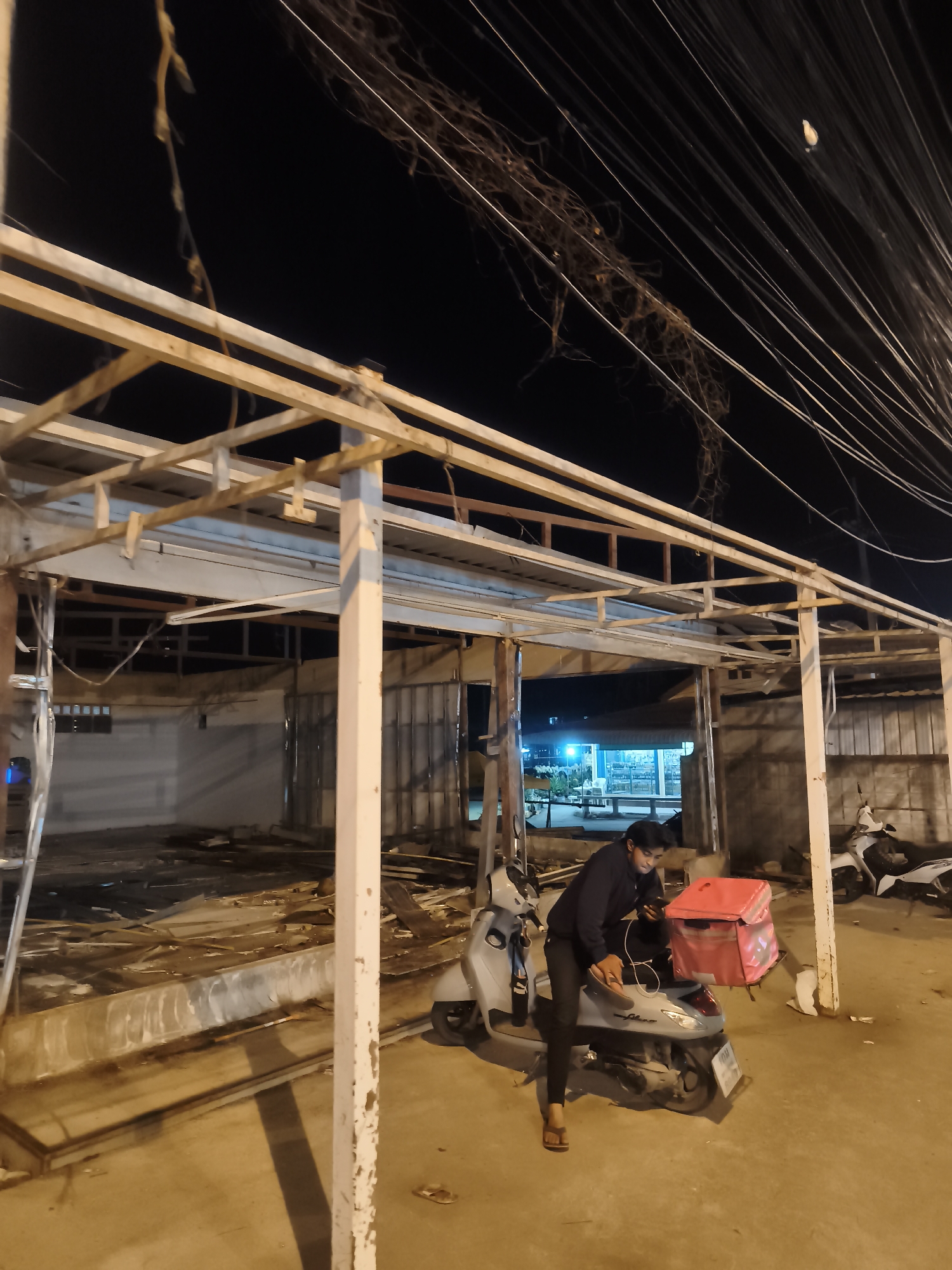 The image shows a man on a scooter, with a red delivery bag, in Baan Tai, Thailand. The man is wearing a black jacket and dark pants. He's seated on a light-blue scooter and appears to be looking at a phone. The scooter is parked near an open structure with a metal frame and roof. The area around the structure is cluttered. Above, a jumble of electrical wires runs across the dark sky. Another scooter is parked further away. The scene appears to be at night or in low light.