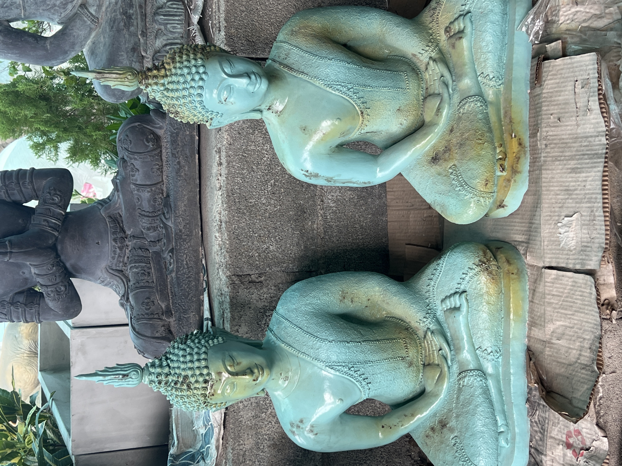 Two teal-colored statues of the Buddha are seated in meditation side-by-side. Both statues are identical, with their hands resting in their laps. They are positioned on a rough-textured stone surface, with the background showing a large, dark, seated figure and some greenery. The statues appear to be made of a material that has a patina, with areas of greenish-blue and yellow-brown discoloration. They are placed on pieces of cardboard. The scene is outdoors, with the light suggesting it is daytime. The location is identified as Ko Pha-ngan District, Thailand.