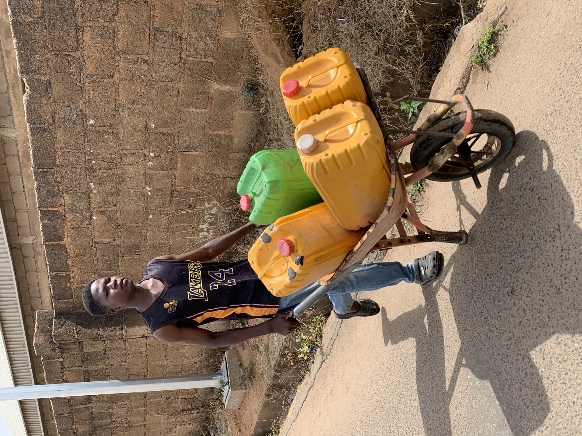 A young man is depicted outdoors, possibly in Jos, Nigeria, based on contextual clues. He is wearing a black and gold Lakers jersey, blue jeans, and black slip-on shoes. He is pulling a heavily loaded wheelbarrow containing three plastic containers, two yellow and one green, each with a colored cap (red or white). The wheelbarrow is rusty. The setting appears to be a street alongside a weathered stone wall. The light suggests it is daytime. His shadow is visible on the pavement.