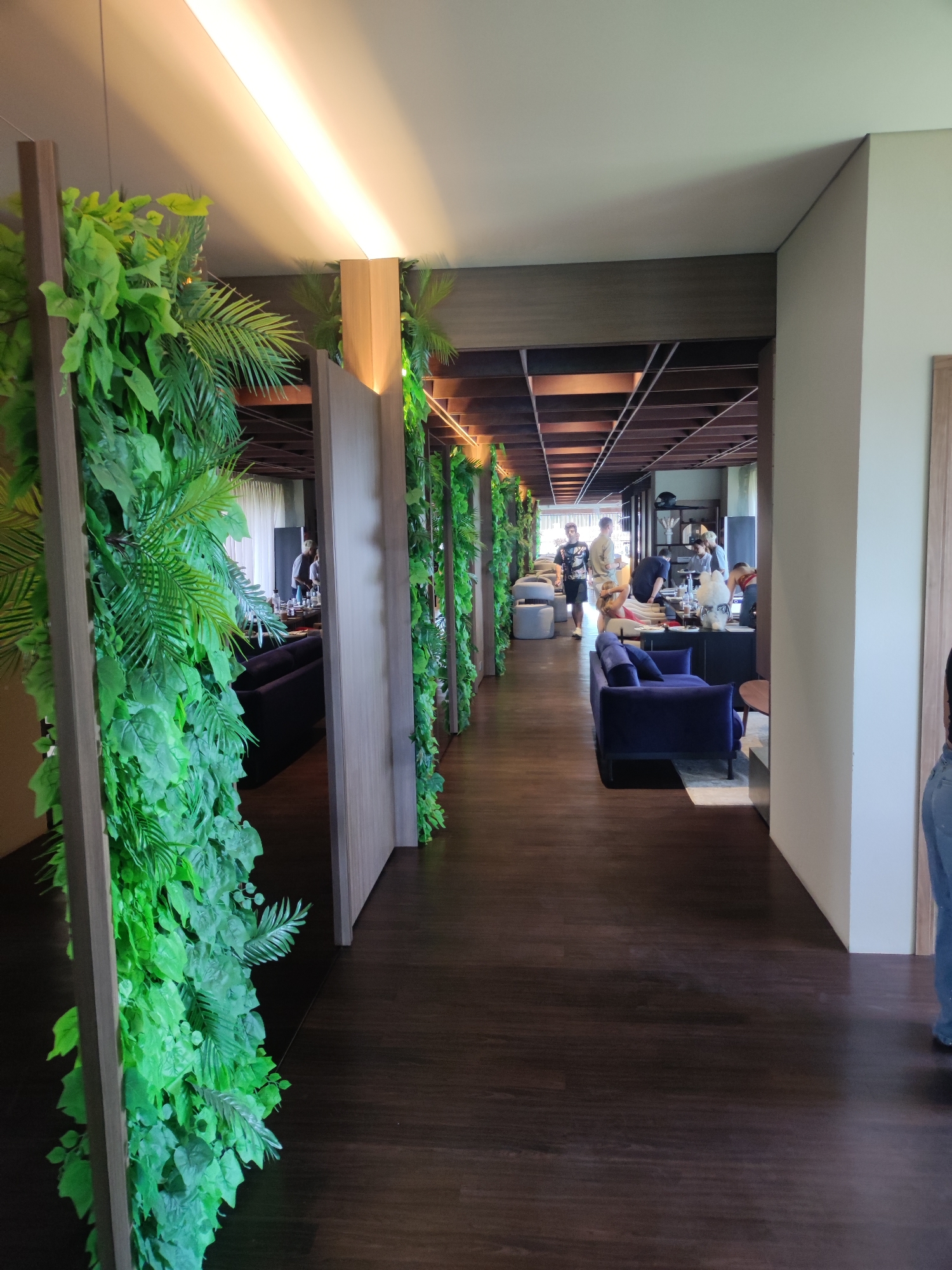 The photograph depicts an interior space with a modern design, featuring a long hallway with a dark wooden floor. Along the hallway, there are vertical structures covered in lush green foliage, creating a garden-like effect. These structures appear to be partitions or screens that partially obscure the view into adjacent rooms.
In the background, through the openings created by the screens, there is a dining or lounge area with dark-colored sofas and chairs. People can be seen in this area, some standing and others seated, suggesting a social gathering or a restaurant setting. The space beyond seems well-lit, with natural light filtering through large windows or openings.
The overall environment is contemporary and stylish, with a focus on integrating nature into the interior design. The use of greenery and the open layout give the space a relaxed and inviting atmosphere.
While the prompt mentions North Kuta, Indonesia, this information is not visually verifiable within the provided image. Therefore, the location cannot be confirmed based solely on the image content.