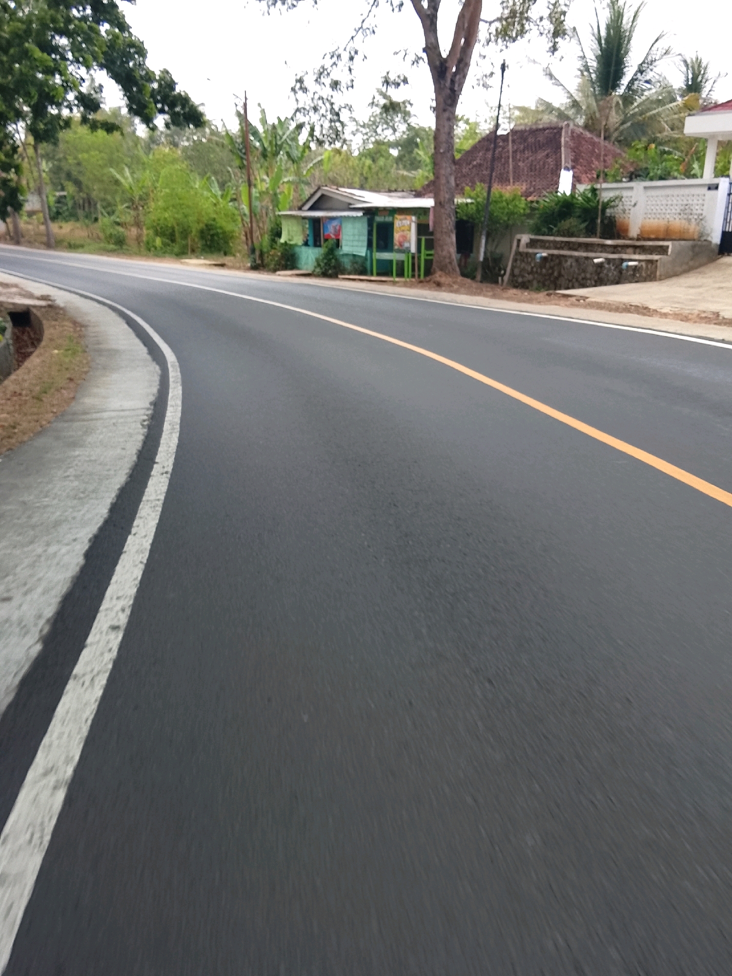 The image depicts a roadside scene in Banjar, Indonesia, featuring an asphalt road with a yellow dividing line and a white edge line. To the left of the road, a concrete drainage ditch runs alongside the asphalt. Further left, vegetation and trees are visible. On the right side of the road, a small, green and blue roadside stall with a white awning is situated. Behind the stall, a traditional Indonesian house with a tiled roof can be seen, along with other trees and a white wall. The sky is overcast. The perspective suggests the image was taken from a moving vehicle, likely a motorcycle or car, due to the motion blur on the road. There are no people or animals clearly visible in the scene.