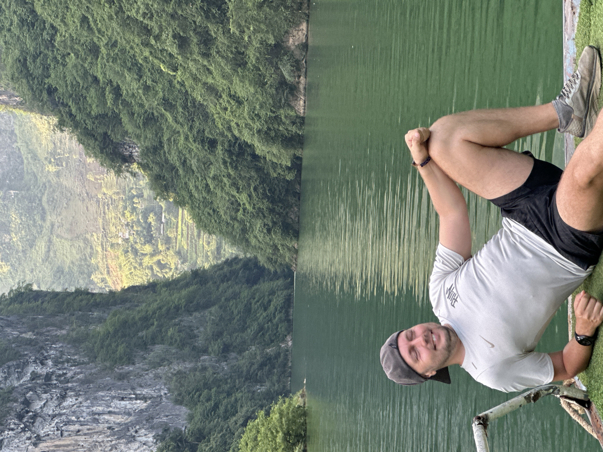 A landscape scene in Đồng Văn Commune, Vietnam, shows a Caucasian male lying on his side along a lake's edge. The man wears a light gray t-shirt, black shorts, a gray baseball cap, and gray running shoes with gray socks. His arms are positioned with his fists clasped together. He wears a watch on his left wrist and a woven bracelet on his right. He is positioned near a white painted metal railing.
To the left of the lake is a verdant mountain covered in lush vegetation, with areas of exposed rock. The lake's water appears green and reflects the surrounding landscape. The overall scene suggests a natural, outdoor setting.