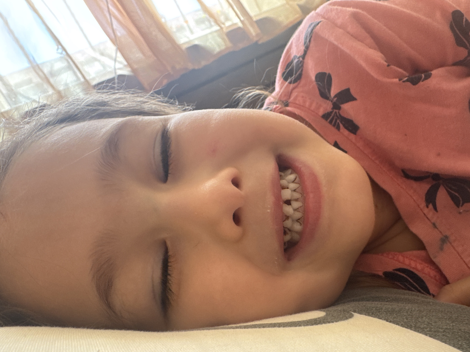 A young girl is lying down with her eyes closed and a partial smile, revealing her teeth. She is wearing a pink long-sleeved shirt with dark floral patterns. She is resting on a pillow or cushion with a white and grey patterned cover. In the background, there is a window with sheer, light-colored curtains. The location is unknown, and no information suggests Tashkent, Uzbekistan.