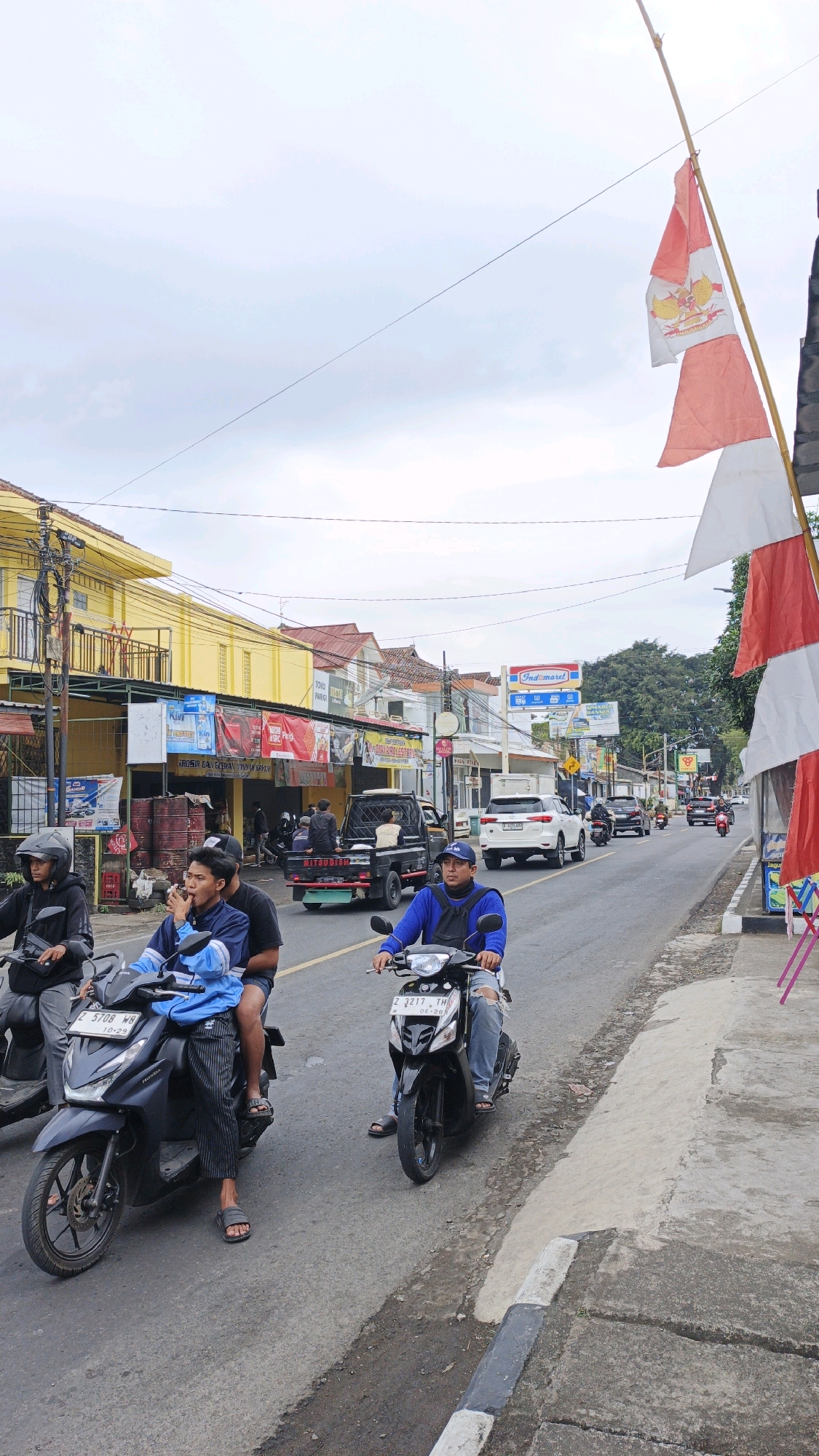 In the village of Desa Karangbenda, Indonesia, a street scene unfolds under an overcast sky. The main focus is a road populated with motorcycles and vehicles. In the foreground, two men ride a black motorcycle; the driver wears a blue jacket and jeans, while the passenger, with a black shirt, sits behind him. To their left, another motorcycle carries two individuals, one of whom is smoking. The road is flanked by buildings, including a yellow structure with shops displaying various signs and banners, some in Indonesian. A white SUV and a black pickup truck are also visible on the road, along with other motorcycles in the distance. Indonesian flags, red and white, are prominently displayed on a bamboo pole on the right side of the image, suggesting a local celebration or event. The environment appears to be a typical street in a Southeast Asian village, with modest buildings, overhead utility lines, and a paved road.