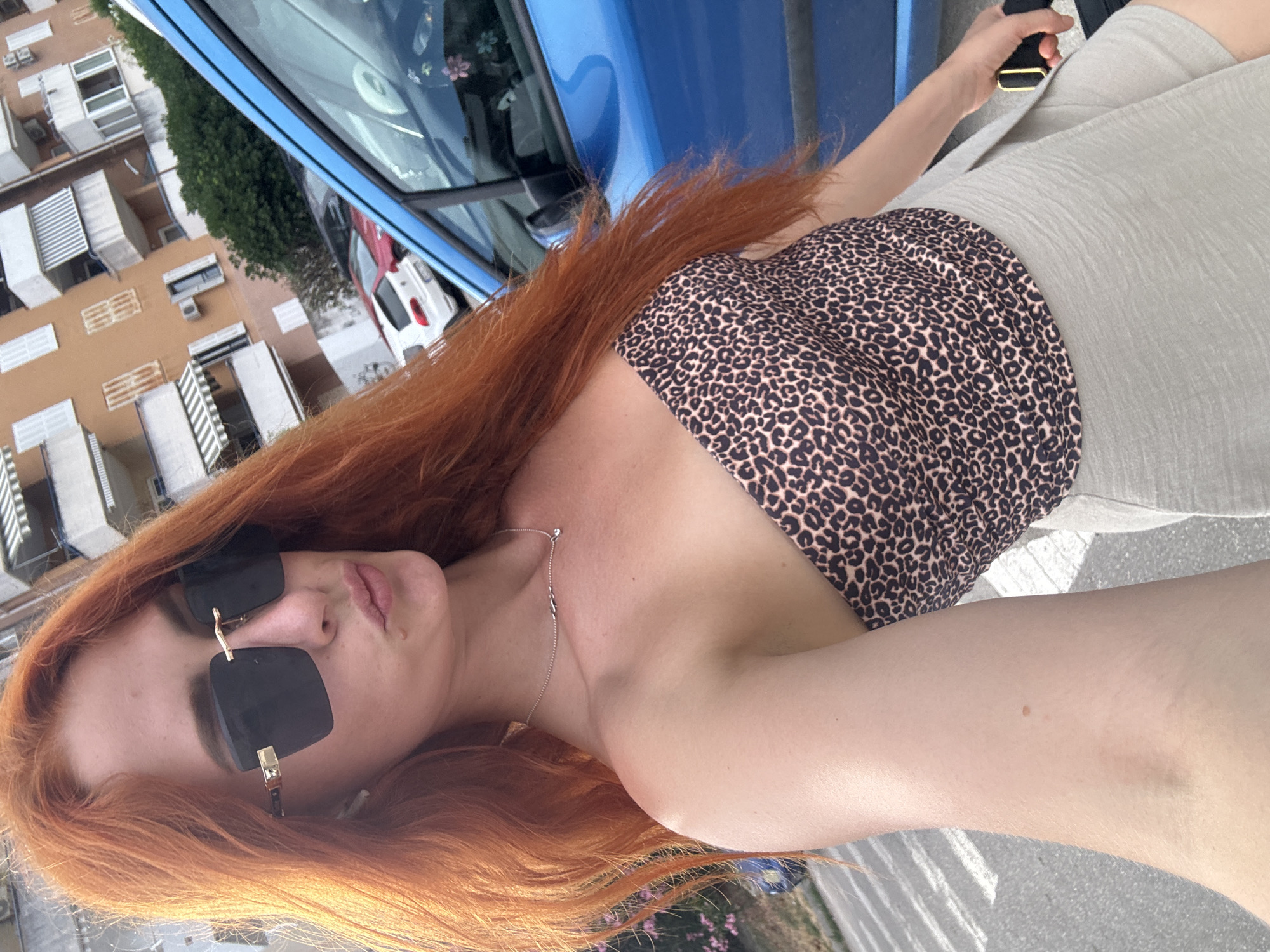 The media file shows a young woman with long, auburn hair, wearing large, black-rimmed sunglasses and a leopard-print halter top. She is wearing a light-colored, textured skirt or shorts. The image is taken outdoors, with a blue car prominently in the background. Buildings, possibly apartments, are visible to the left, suggesting an urban or suburban setting. The woman is holding a black object with a gold accent in her right hand, which appears to be a phone or similar device.
The angle of the shot suggests the photo was taken by the woman herself, possibly a selfie. The lighting indicates it is daytime, and the scene appears to be an outdoor area with pavement. The overall mood is casual and relaxed. There is no visible text in the image. The location context provided is Budva, Montenegro.
