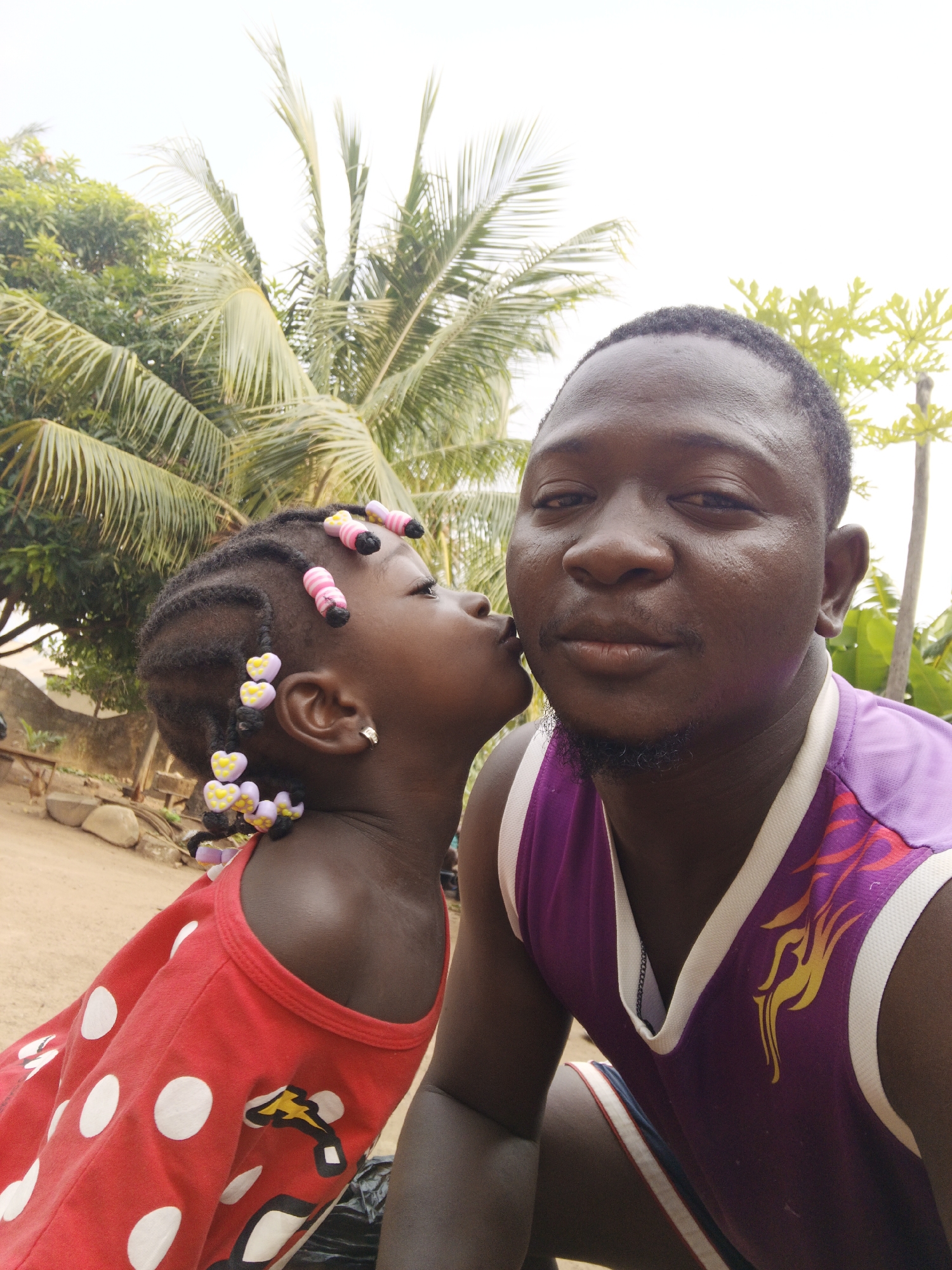 A close-up photo shows a man and a young child in an outdoor setting. The man is positioned to the right, and the child is on the left, kissing him on the cheek.
The child, a young girl, has braided hair adorned with colorful beads. She wears a red shirt with white polka dots.
The man has short black hair and a closely trimmed beard. He wears a purple sleeveless shirt with white trim and a flame design.
In the background are green trees, including a palm tree. Based on this image, the precise location in Akwanga, Nigeria cannot be determined.