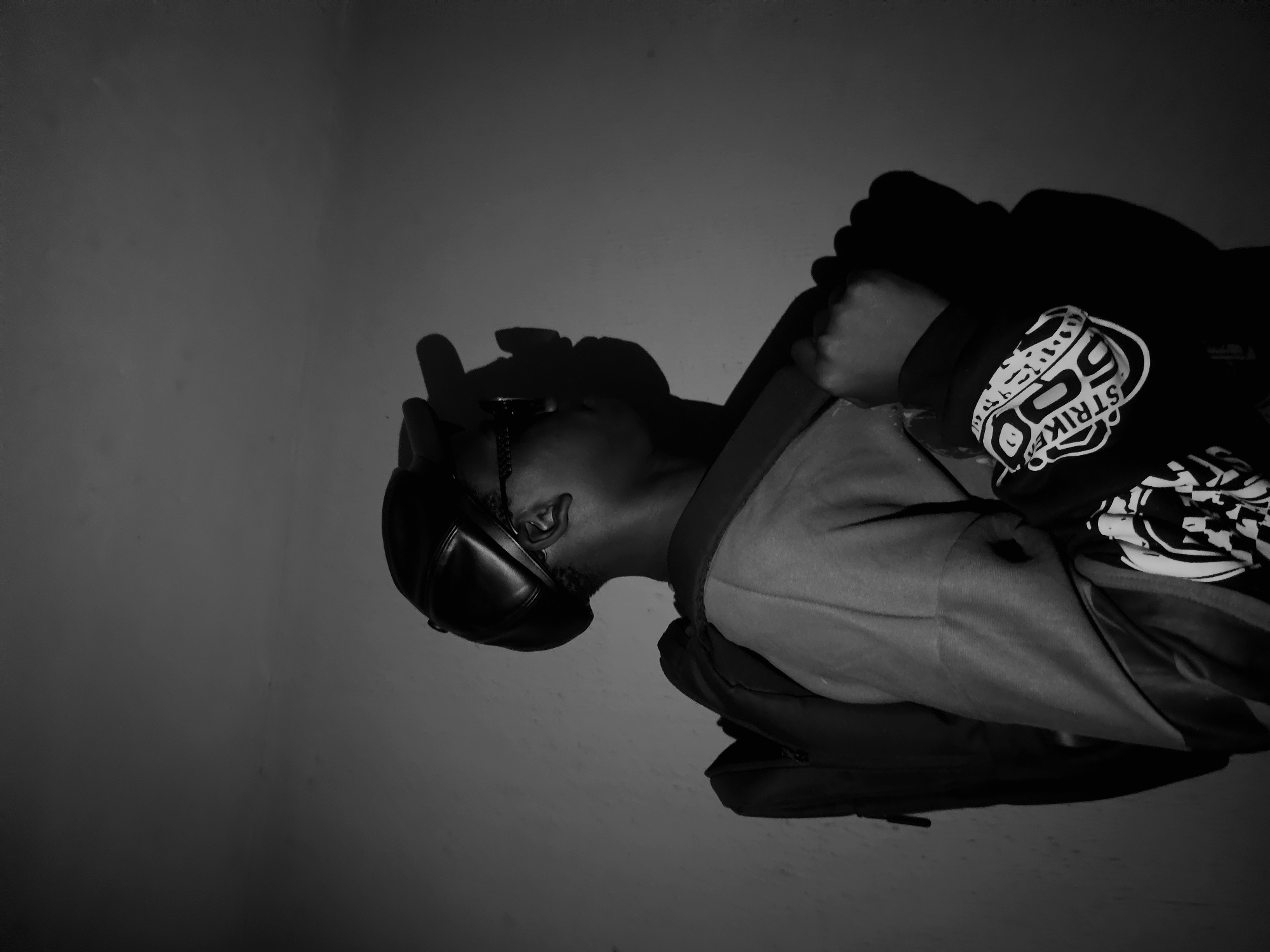 A black and white photograph shows a man wearing a cap, jacket, and hoodie. The man is positioned in the lower half of the frame, facing the left side, with the left side of the image in focus. The man’s cap is dark, and his jacket is partially visible over a light shirt. The hoodie has a white design that says "Strike". The background of the photo consists of a plain, dark wall. There are no other identifiable elements in the photograph that reveal information about the city of Nkwelle, Nigeria.