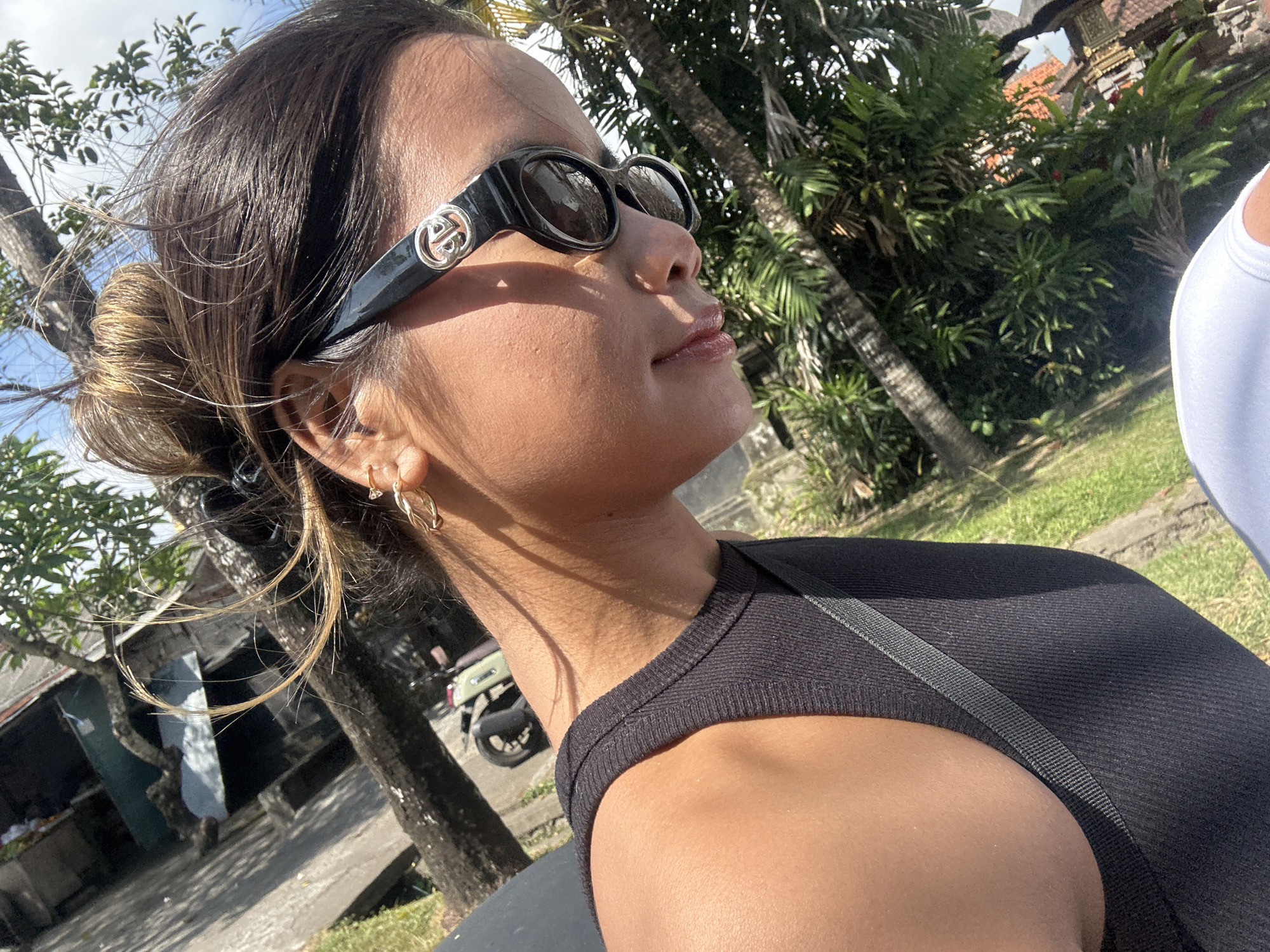 An eye-level, medium shot captures a young woman wearing a black tank top and black sunglasses with a silver logo on the right lens. Her brown hair is pulled up in a messy bun, with several strands falling around her face and neck. She has on a small hoop earring in her left ear. The woman is looking down and to the right, with her face angled away from the camera. The background is a blur of greenery, suggesting an outdoor setting, possibly in a tropical climate. The image appears to be taken on a sunny day, with strong shadows visible. Based on the distinctive architecture and vegetation visible in the background, the location is likely Sukawati, Indonesia.