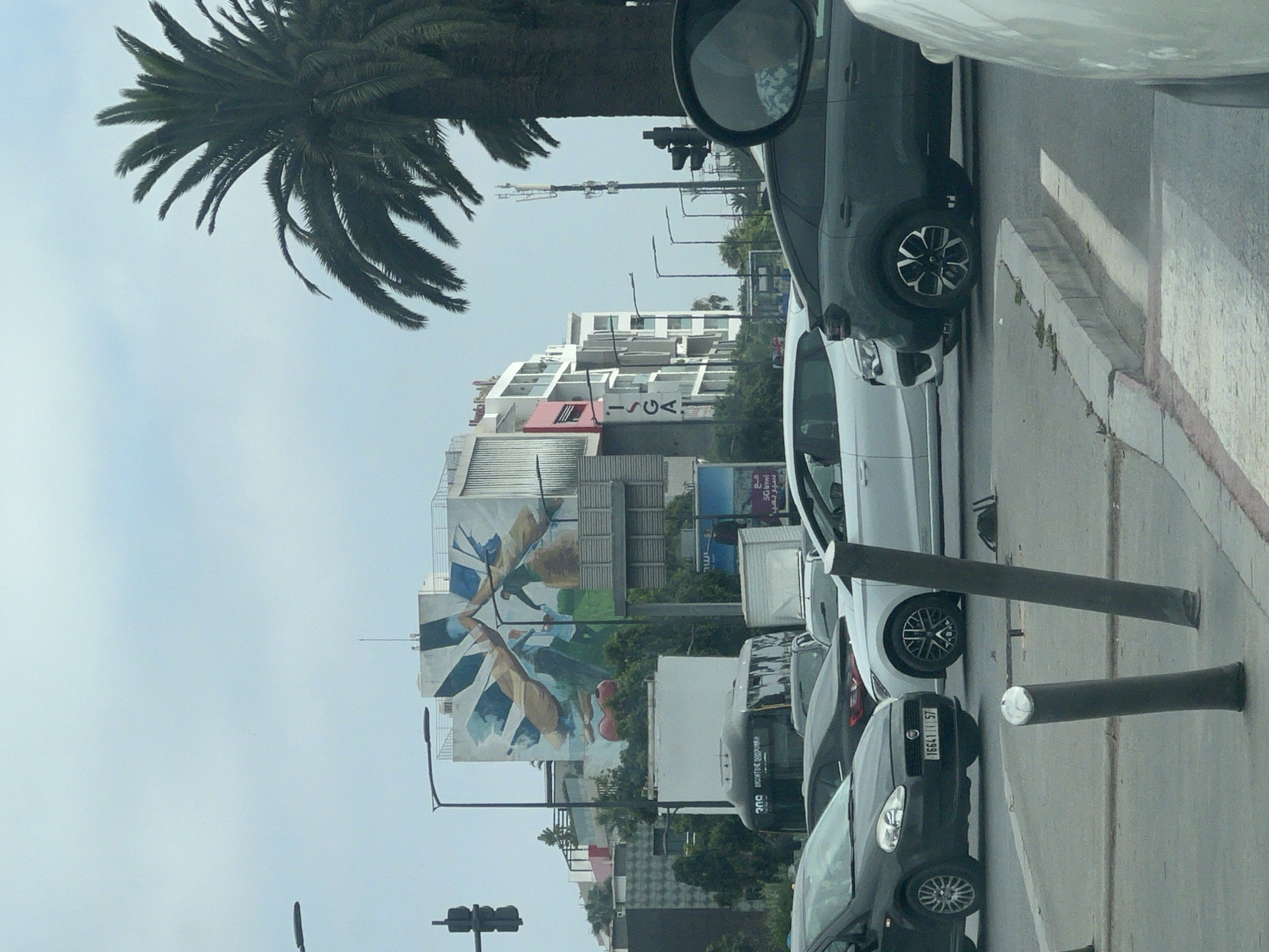 The image depicts an urban street scene under an overcast sky, identified as Casablanca, Morocco, by the visible "ISGA" building and a license plate (16641 | 57) consistent with the region. Multiple vehicles are present, suggesting traffic. In the left midground, a dark grey Fiat Punto with the aforementioned license plate is prominently featured, facing right. Behind it, a dark sedan is partially visible. To the right of the Fiat, a white car's front section is seen. In the right midground, a dark grey crossover SUV with distinctive wheel rims is stopped. The extreme right foreground shows part of a light-colored vehicle, and its side-view mirror reflects a passenger wearing a patterned shirt. Two dark grey cylindrical bollards, one with a reflective top, lean slightly on the pavement in the immediate foreground.
In the background, multiple buildings line the street. On the left, a multi-story building with white and grey facades features "ISGA" written vertically and has balconies with red accents. Adjacent to it, a large building displays a prominent, colorful wall mural depicting abstract human-like figures interacting with large, flowing fabric-like elements and incorporating violin shapes at the bottom, predominantly in shades of blue, brown, green, and white. A black public transit bus, marked with "ona" and Arabic script, is visible behind the mural building. Further right, a series of billboards include one displaying Arabic text and "5G" and "6G" logos. Tall palm trees are visible on the right side of the image, partially obscuring a cellular antenna tower. Several streetlights and traffic light poles are interspersed throughout the scene.