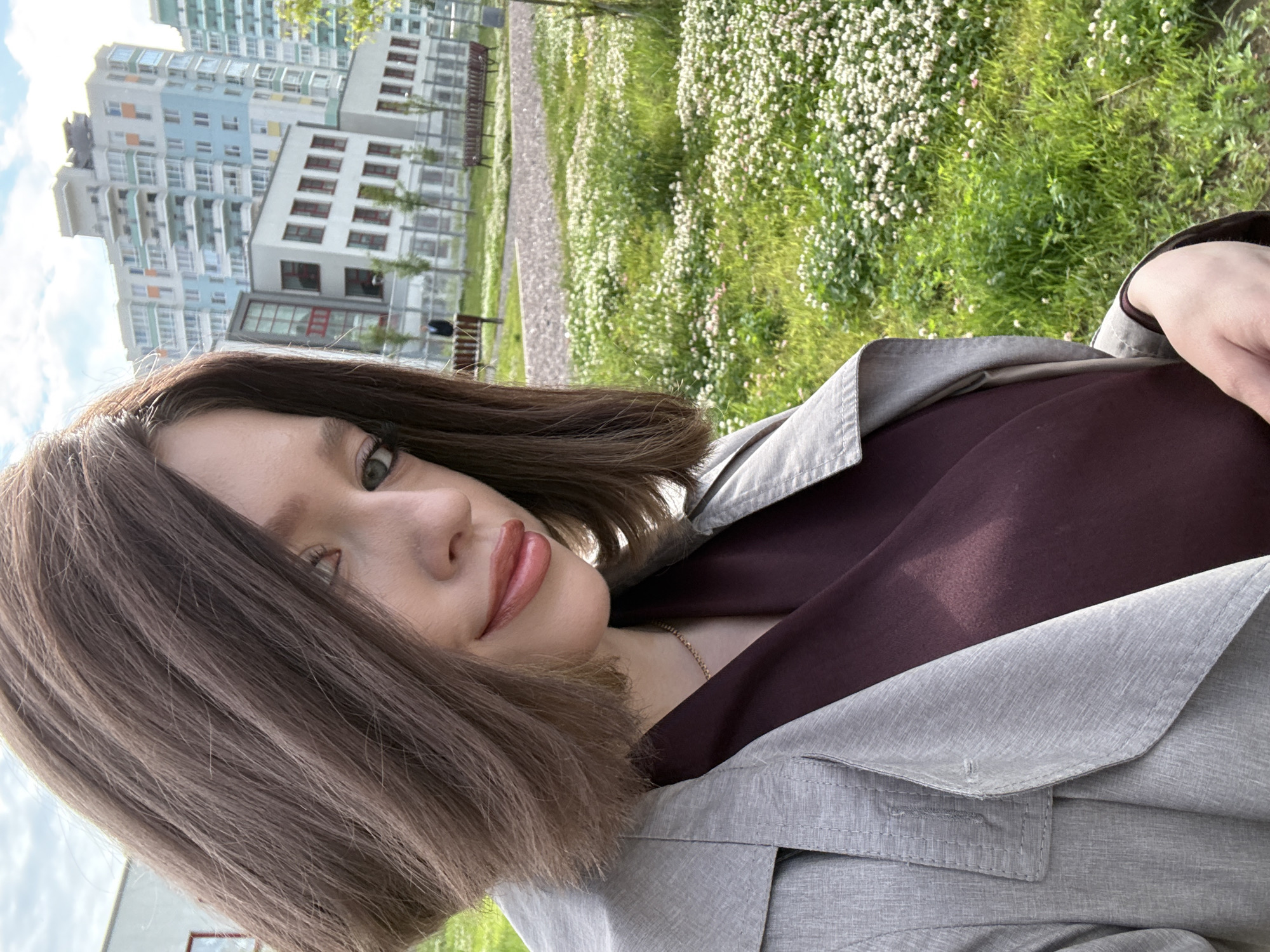The image shows a young woman with shoulder-length brown hair, wearing a light gray trench coat over a dark plum-colored garment. She is taking a selfie outdoors in what appears to be a modern urban park. Behind her, several multi-story residential buildings with colorful facades are visible, along with benches and grassy areas with small white flowers. The presence of these buildings and the style of architecture are consistent with residential areas in Saint Petersburg, Russia, particularly newer developments. The lighting suggests it is daytime, possibly late afternoon given the angle of the sunlight.