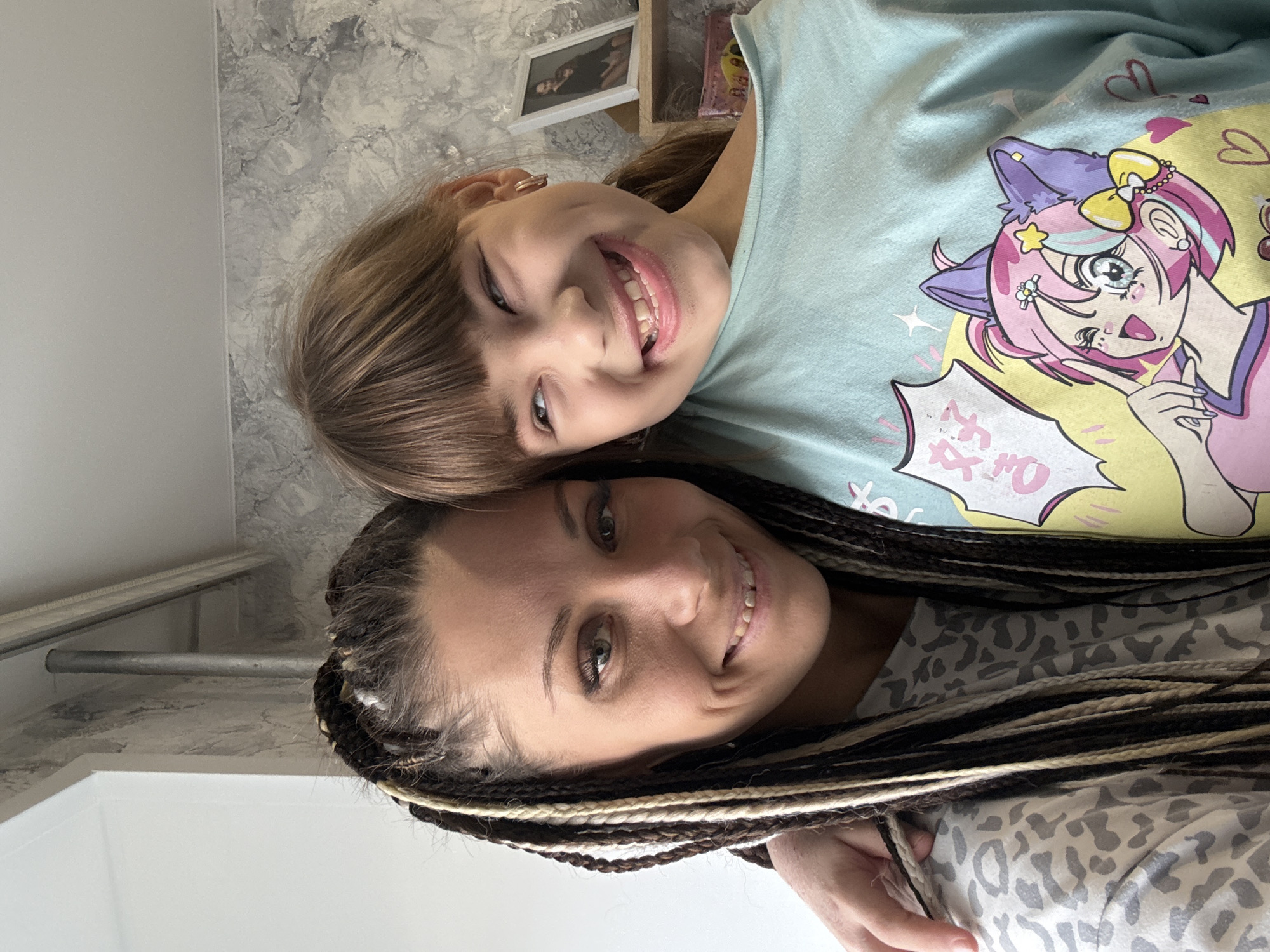 The image shows two smiling individuals: a young girl and a woman. The girl has shoulder-length brown hair with bangs and is wearing a light blue t-shirt with an anime-style graphic. The woman has dark hair styled in braids with blonde strands. They are positioned closely together, possibly embracing. The background includes a wall with a textured, marble-like pattern. In the upper part of the image, framed pictures and wooden shelves are visible. The location cannot be determined to be in Rusanovo, Russia from the given image.
