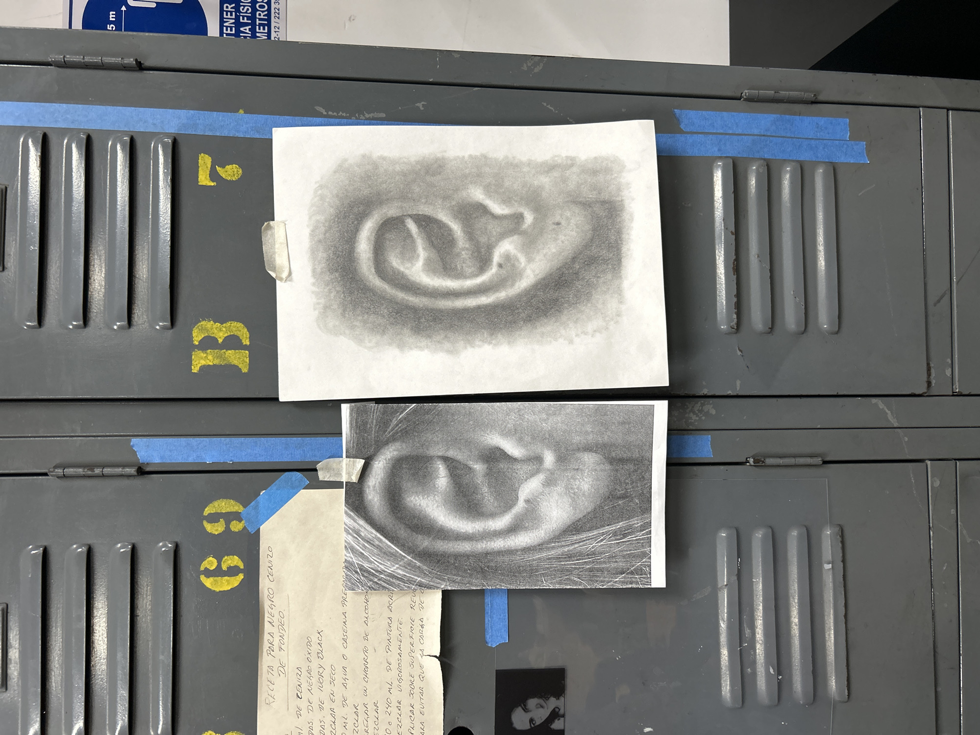 This image captures a close-up view of a gray metal locker, possibly in an art studio, with a focus on two drawings of ears taped to the surface. The lockers are marked with yellow stenciled numbers "13" and "69". The drawings are in charcoal or pencil, one with a softer, more blurred shading, and the other with more defined lines and details.
Attached below the bottom drawing, there is a white page of handwritten notes, probably in Spanish, titled "RECETA PARA NEGRO CENIZO DE FONDEO" (Recipe for ash-gray base). A small black and white photograph is also taped below the notes.
Blue painter's tape is used to adhere the drawings and papers to the lockers. The environment appears to be an indoor space, likely an art classroom or studio.
The location cannot be determined solely from this image. The OCR text mentions a phone number that potentially starts with "2-12", which might offer a geographical hint, but it is not sufficient to pinpoint the location to San Andrés Cholula, Mexico, without additional information.